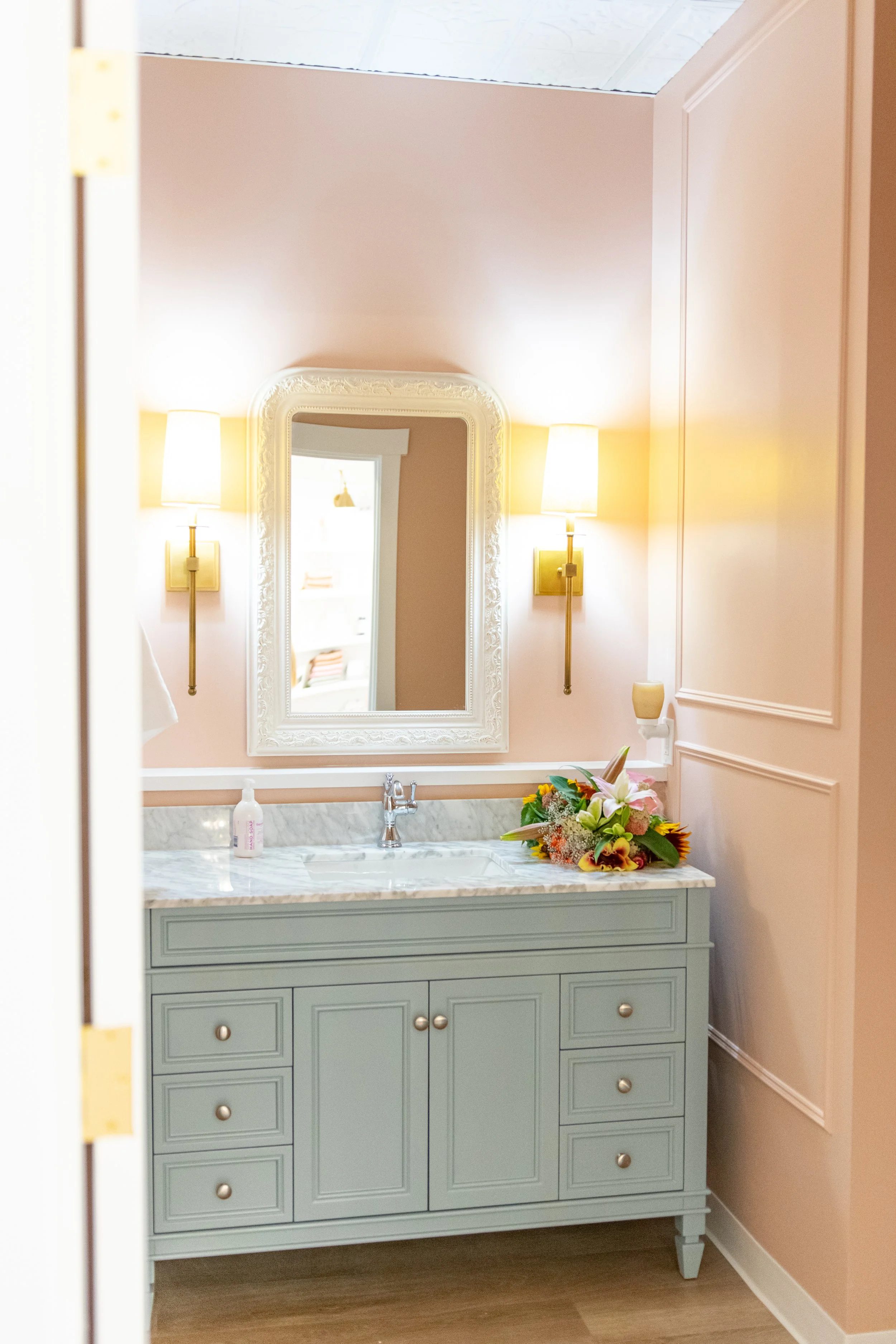 A bathroom vanity with a mirror, two wall-mounted lamps, a pink vase with flowers, and a bottle of soap.