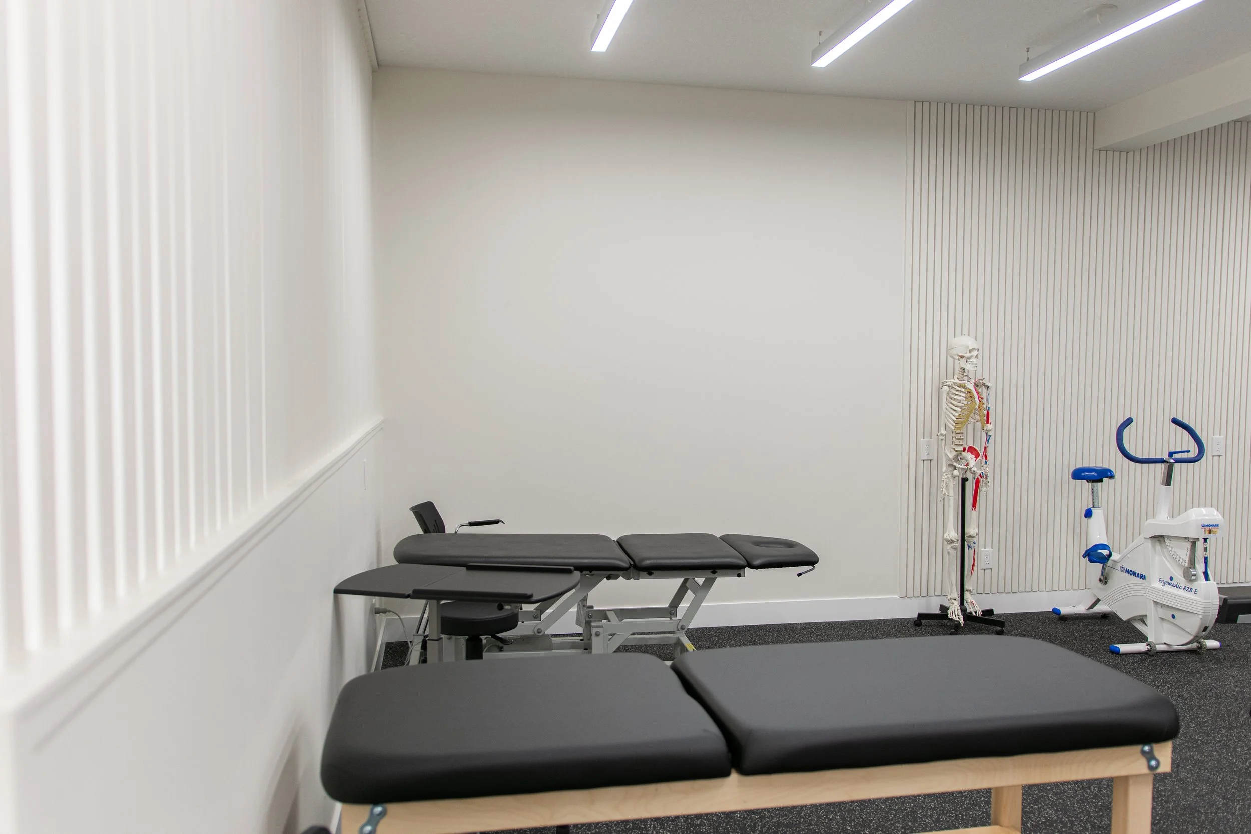 Empty physical therapy or chiropractic treatment room with treatment table, exercise bike, and skeleton model.