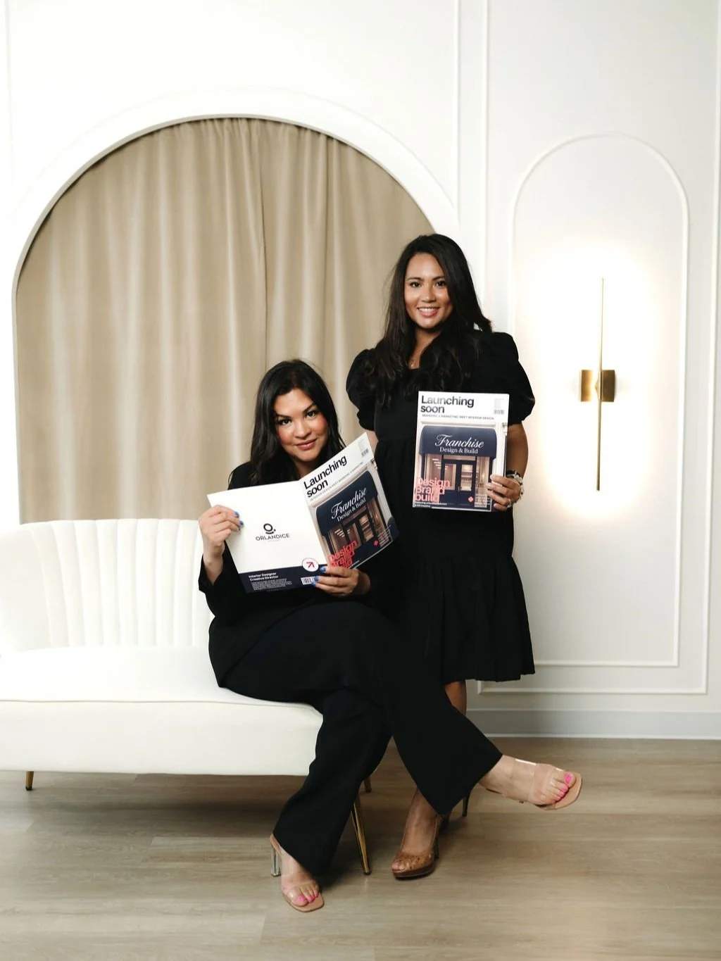 Two women in black dresses holding magazines in a modern, neutral-colored room with a beige curtain and white wall.