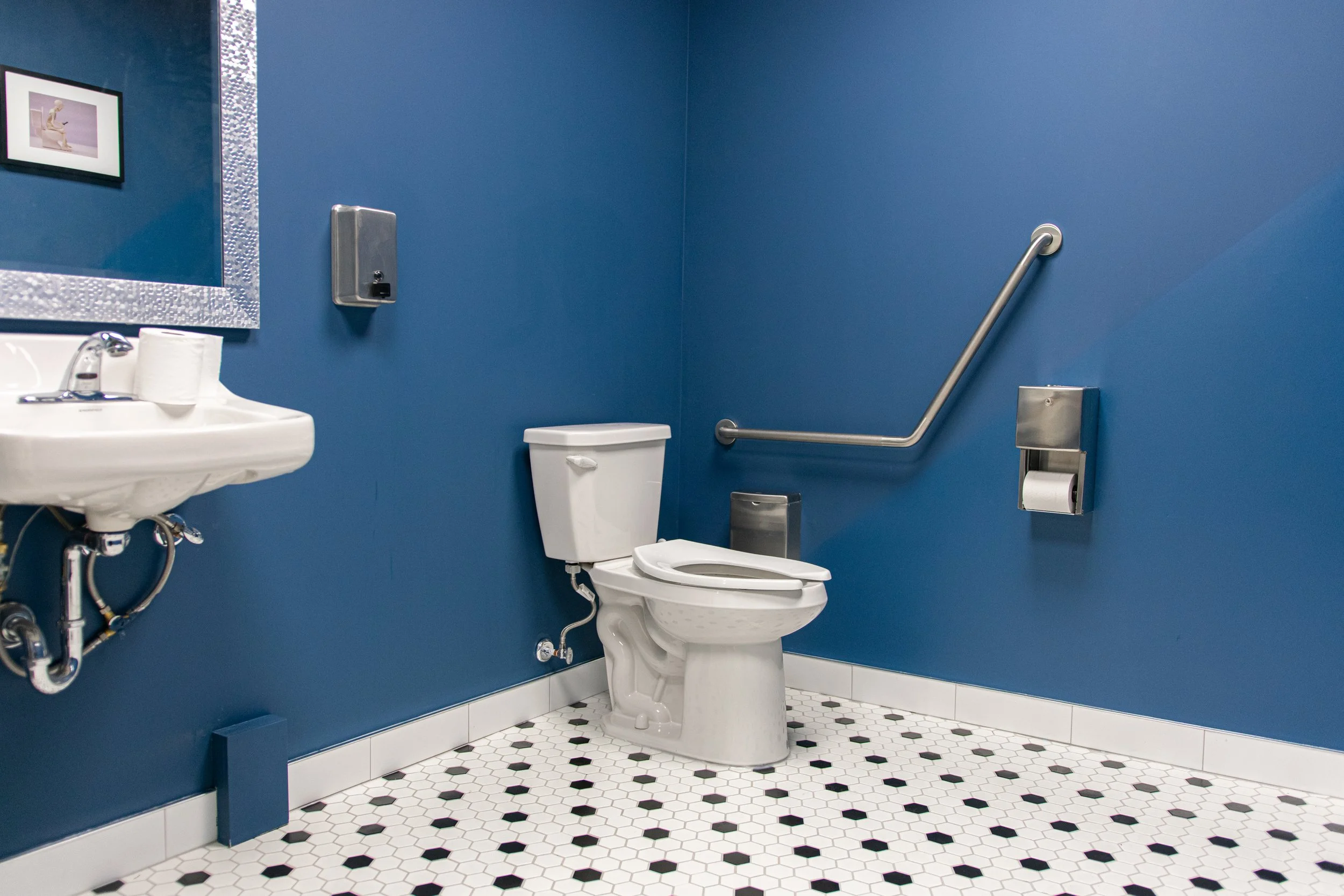 Accessible bathroom with blue walls, a white toilet, a small sink, a mirror with a textured silver frame, and grab bars for support.