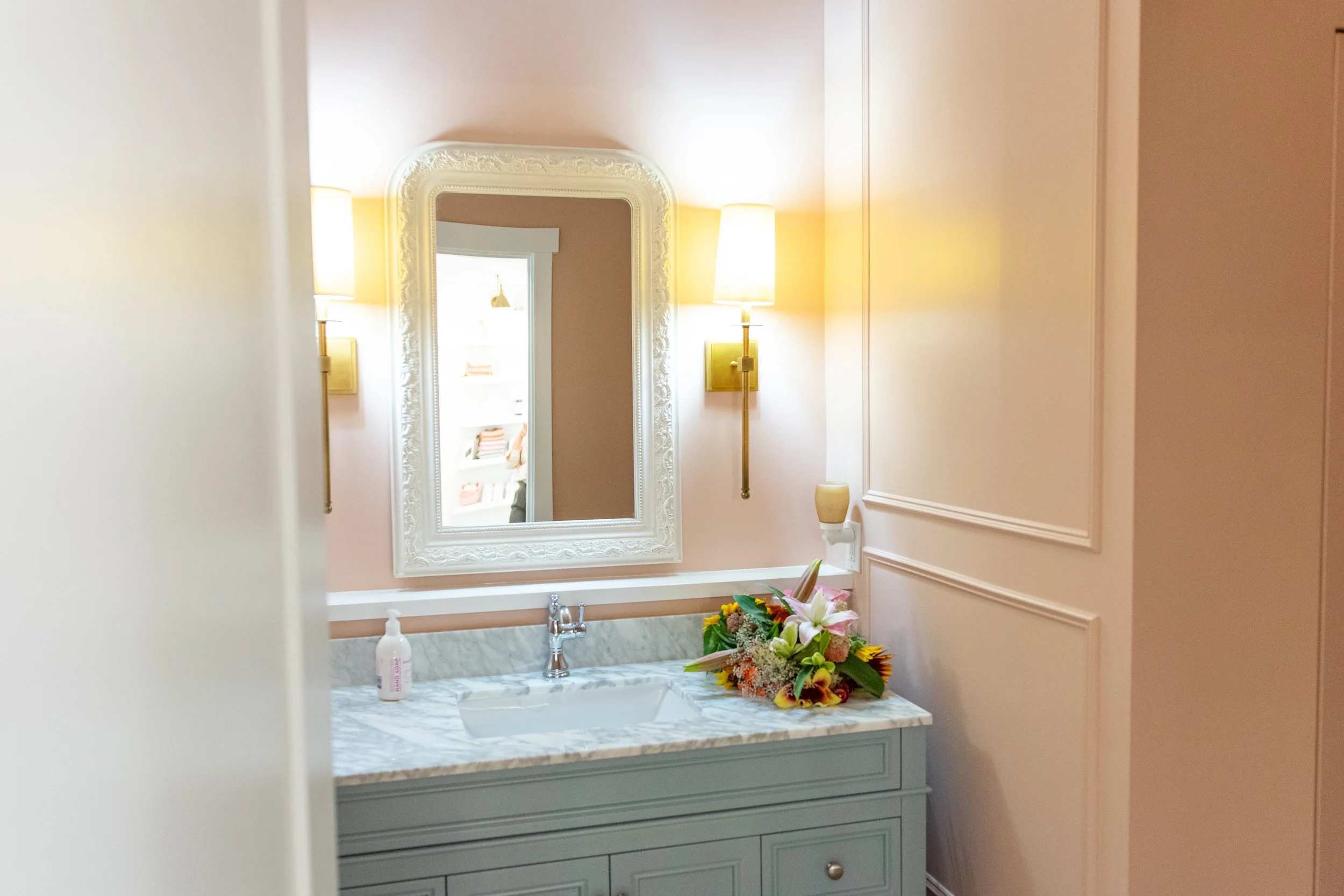 Bathroom with a marble countertop, mirror, wall-mounted lights, pink walls, and a bouquet of flowers.