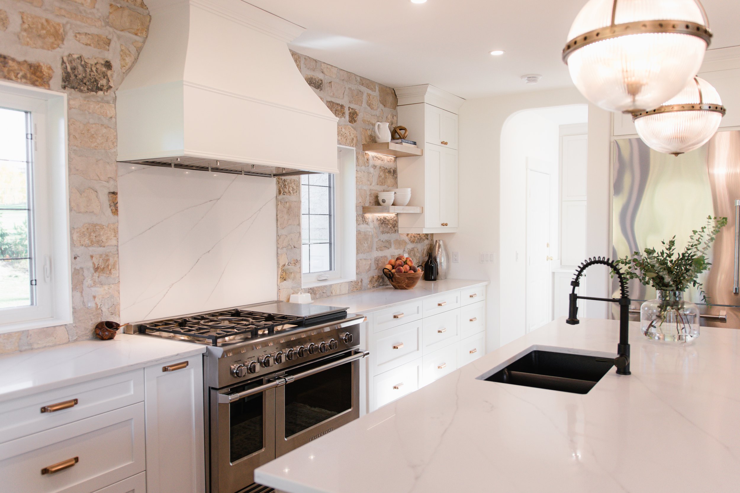 Modern kitchen with white cabinets, marble countertops, and a stainless steel stove. Exposed brick wall behind the stove, with two small windows. Hanging glass light fixtures, a black faucet above a dark sink, and a vase with greenery on the island.