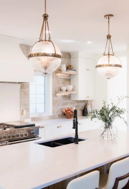 Modern kitchen with white cabinets, stone walls, hanging pendant lights, a window, a black kitchen sink, and a glass vase with greenery on the counter.