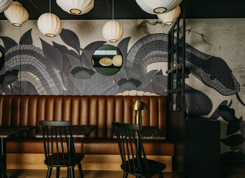Interior of a stylish dining area with a brown leather booth, black chairs, a black partition, and hanging paper lantern lights. A mural of leaves and a snake decorates the wall.