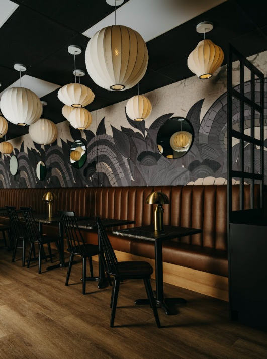 Interior of a modern restaurant or cafe with hanging paper lantern lights, a leather booth, black chairs, and gold table lamps, with a decorative wall mural and a round mirror.
