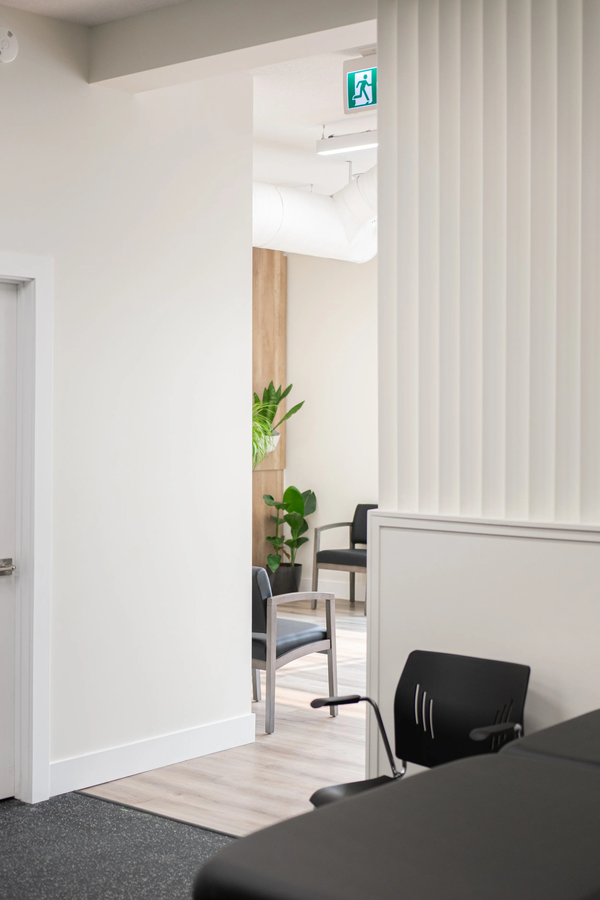 An indoor waiting area with black chairs, potted plants, and a white textured wall. There is a white door on the left and a green emergency exit sign overhead.