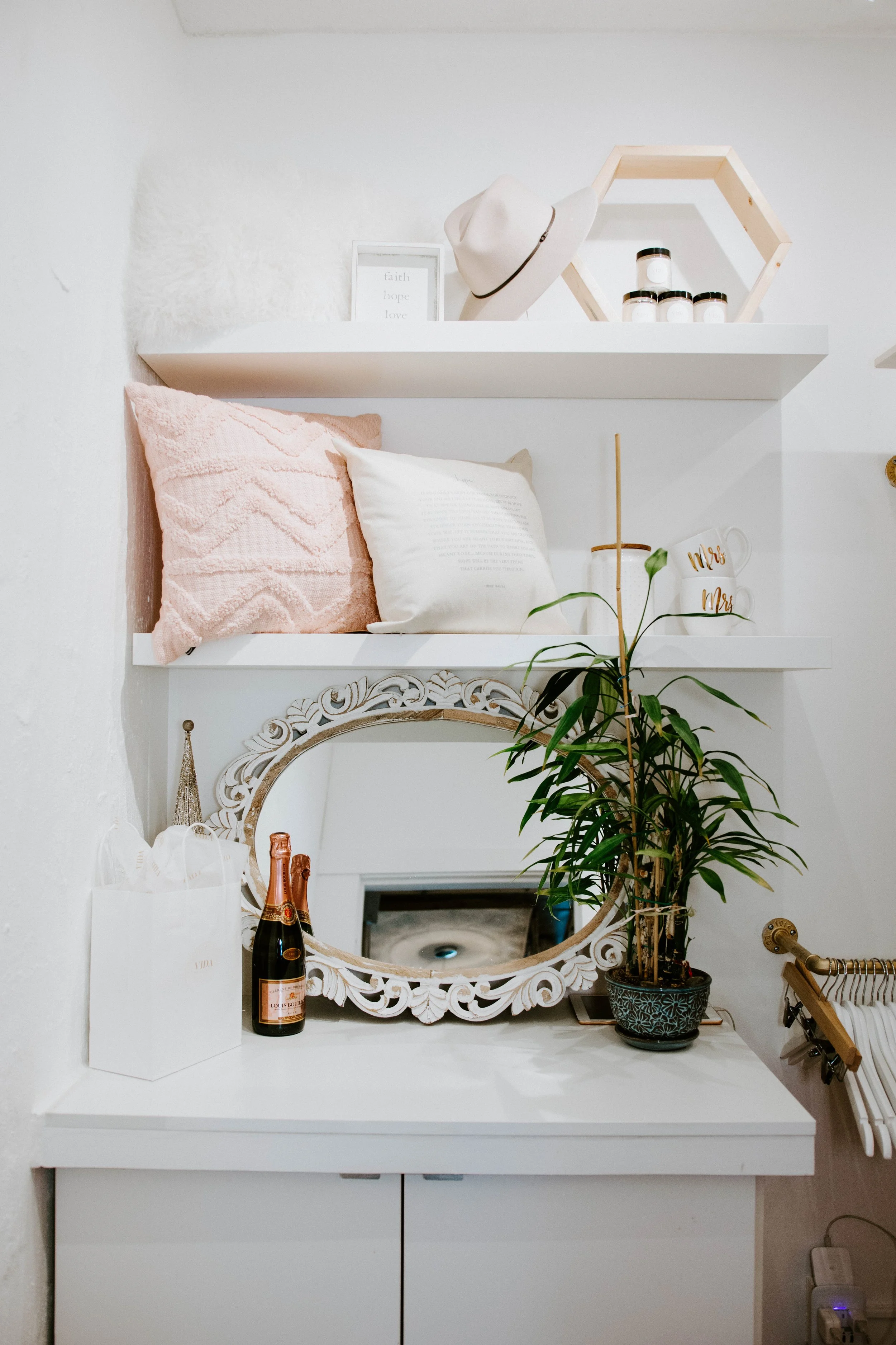 Decorative shelf with pillows, framed quote, hat, honey jars, and 'Mrs' mugs, mirror, champagne bottle, plant, and gift bag on a white cabinet.