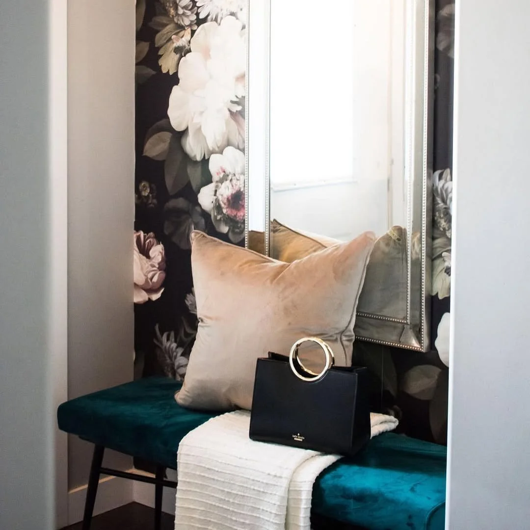 A cozy corner with a plush beige pillow, a black handbag with a circular handle, a white throw blanket, and a teal velvet bench in front of a wall with floral wallpaper, a mirror, and a window.