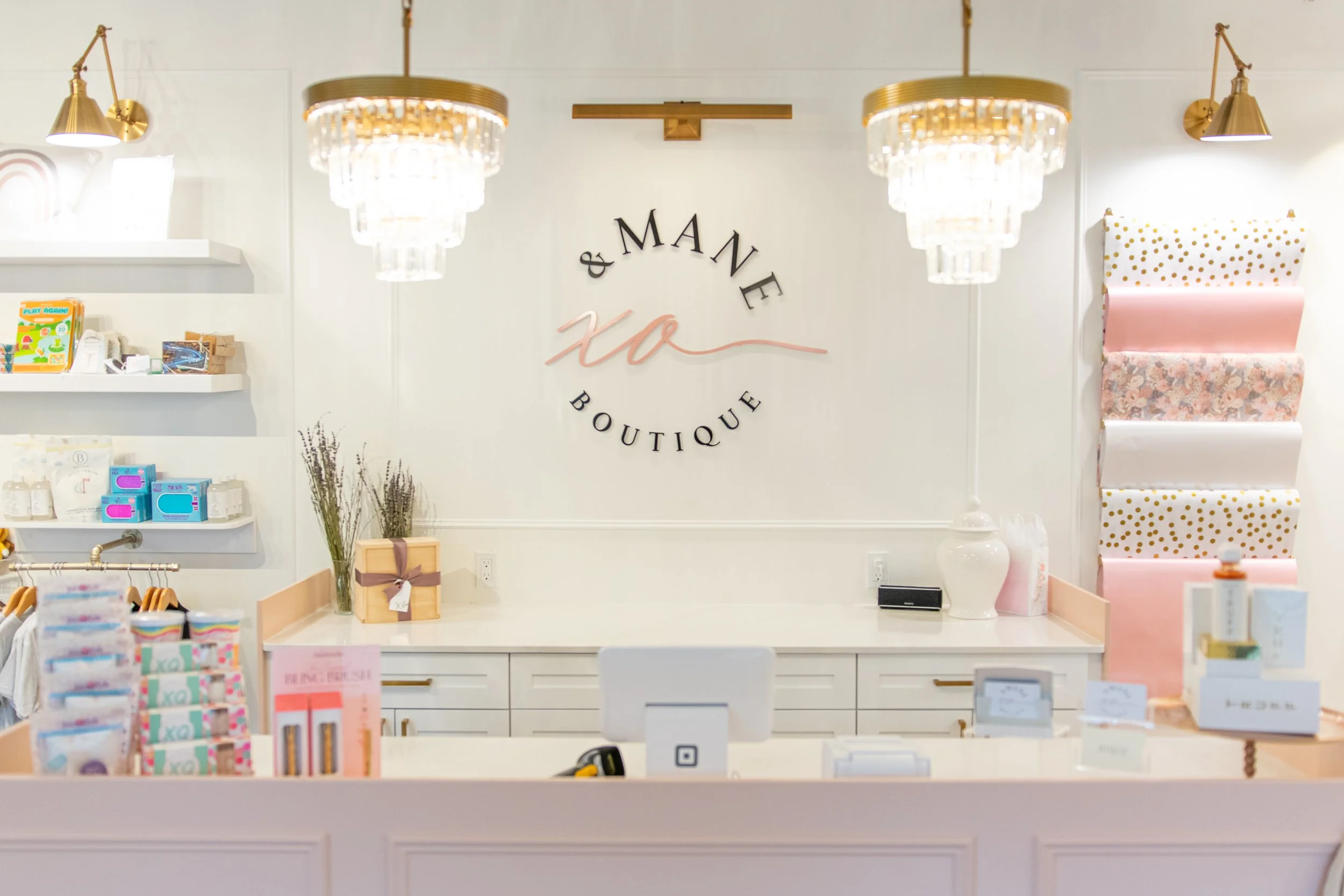 Interior of a boutique shop with white decor, gold accents, hanging chandeliers, shelves with baby and kids' products, a cash register, and a wall sign reading '& M A N E X O Boutique'.