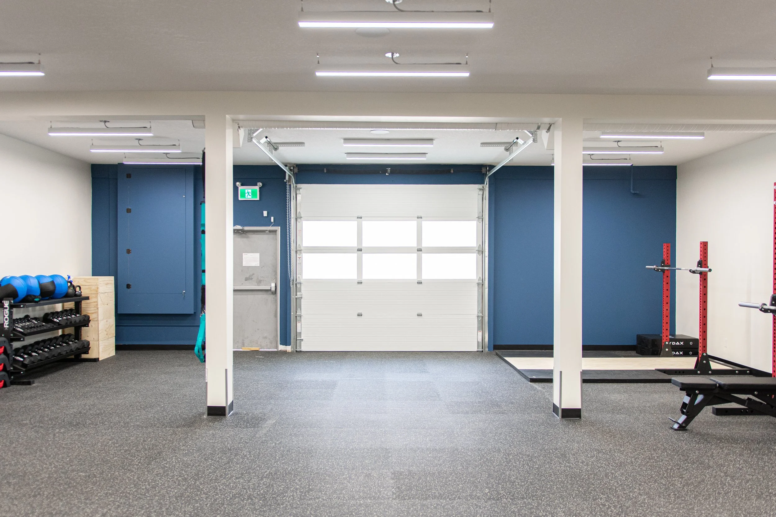 Empty gym with workout equipment including kettlebells, dumbbells, a bench, and squat racks, with a white garage door at the back and blue walls.
