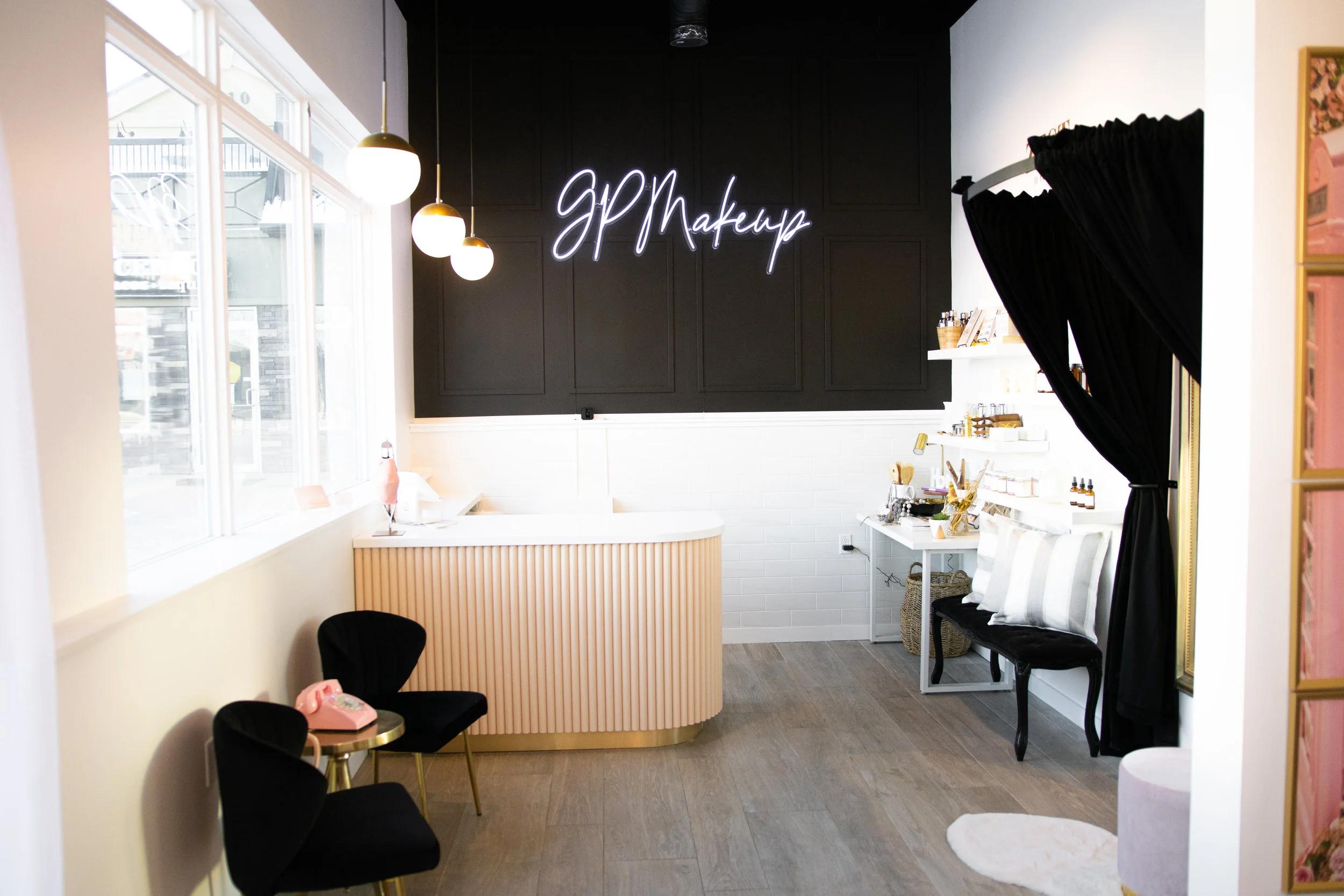 Interior scene of a beauty or makeup salon with a black wall featuring a neon sign that reads 'GP Makeup'. There is a curved reception desk in front of the wall, decorated with vertical beige panels. To the right, there are shelves with bottles and beauty supplies, along with a small table holding makeup brushes and tools. Black curtains are tied back, and a small seating area with black chairs and a pink rotary phone is nearby. Large windows on the left provide natural light, with some hanging globe pendant lights illuminating the space.