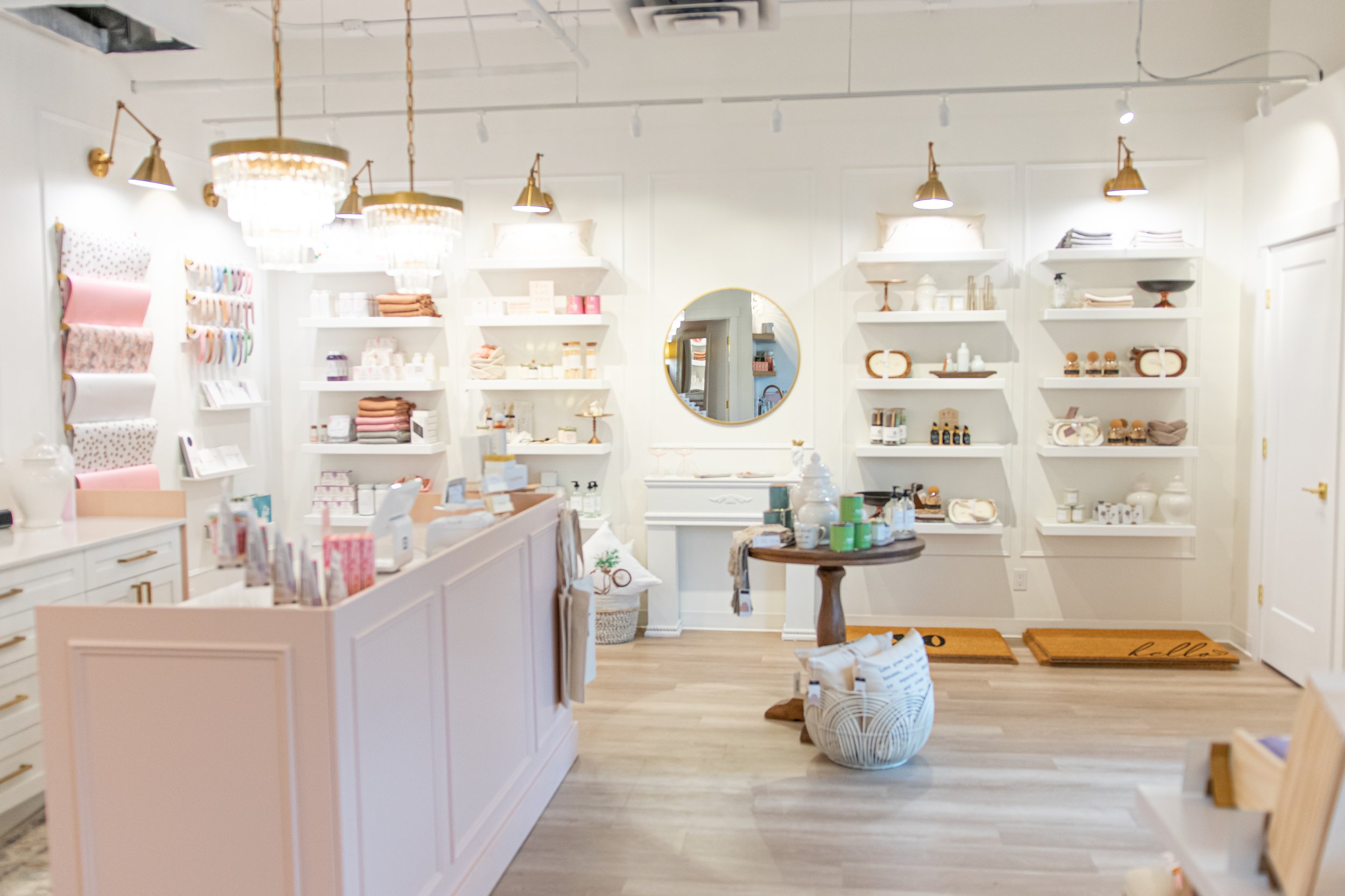 Interior of a retail store with white walls and shelves, displaying home decor items like vases, candles, and towels. There's a peach-colored checkout counter on the left, a round mirror on the back wall, and wooden accents throughout.