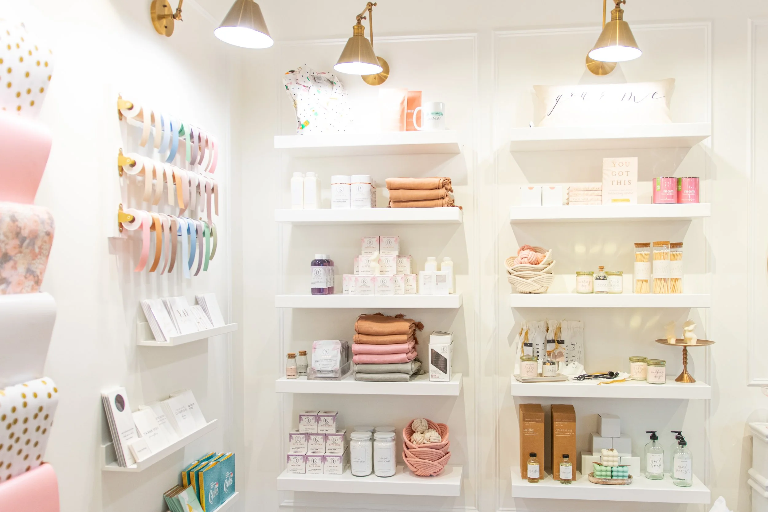 White retail display with shelves filled with candles, soap, skincare products, and decorative items; the left wall features colorful ribbon tags hanging.