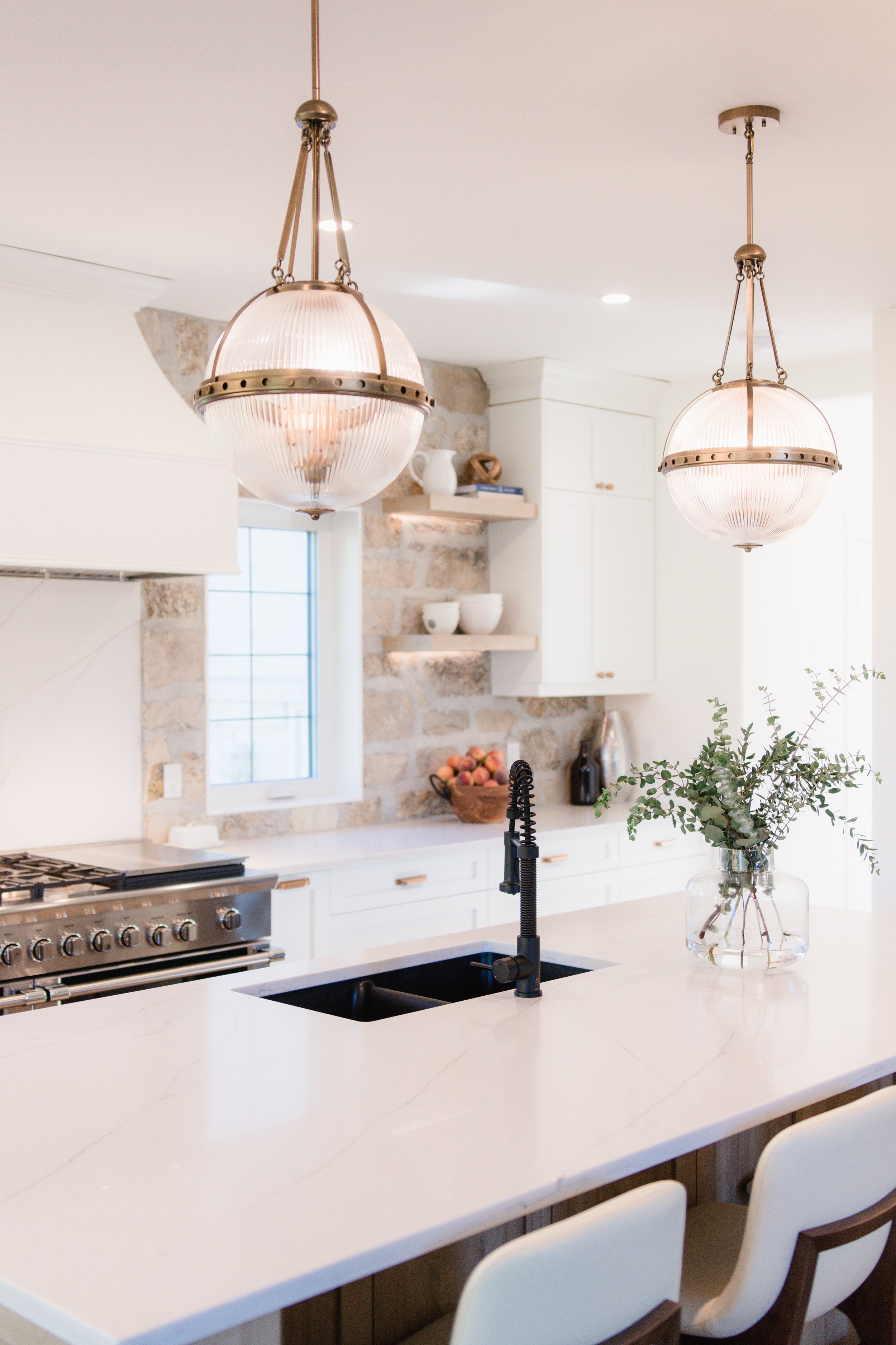 Modern kitchen with white marble island, black sink and faucet, hanging pendant lights, white cabinetry, window, stone accent wall, and a vase with greenery.