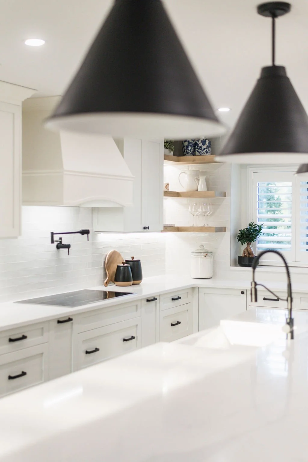 A modern white kitchen with black cabinet handles, open wooden shelves with glassware and decorative items, black pendant lights, and a window with white shutters, a potted plant on the windowsill, and a white countertop with a faucet.