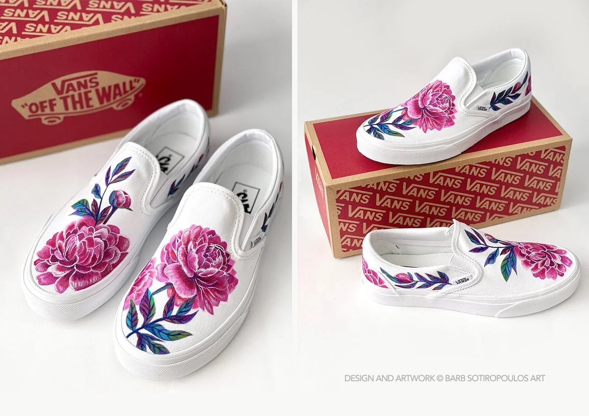 A pair of white Vans slip-on shoes with pink floral artwork and green leaves painted on them, displayed next to their Vans 'Off The Wall' branded shoebox.