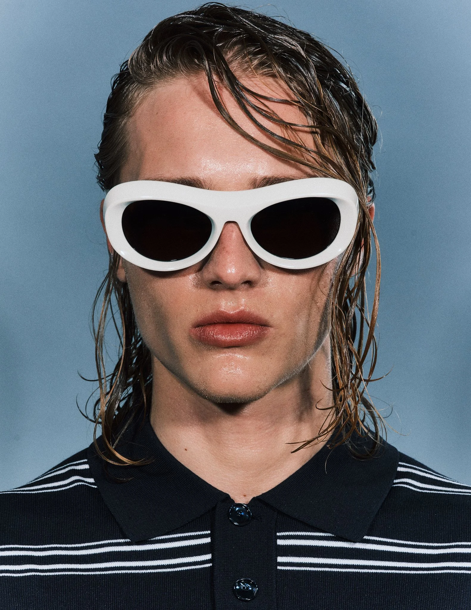 A woman with wet, slicked-back hair wearing oversized white sunglasses and a black and white striped polo shirt, against a plain blue background.