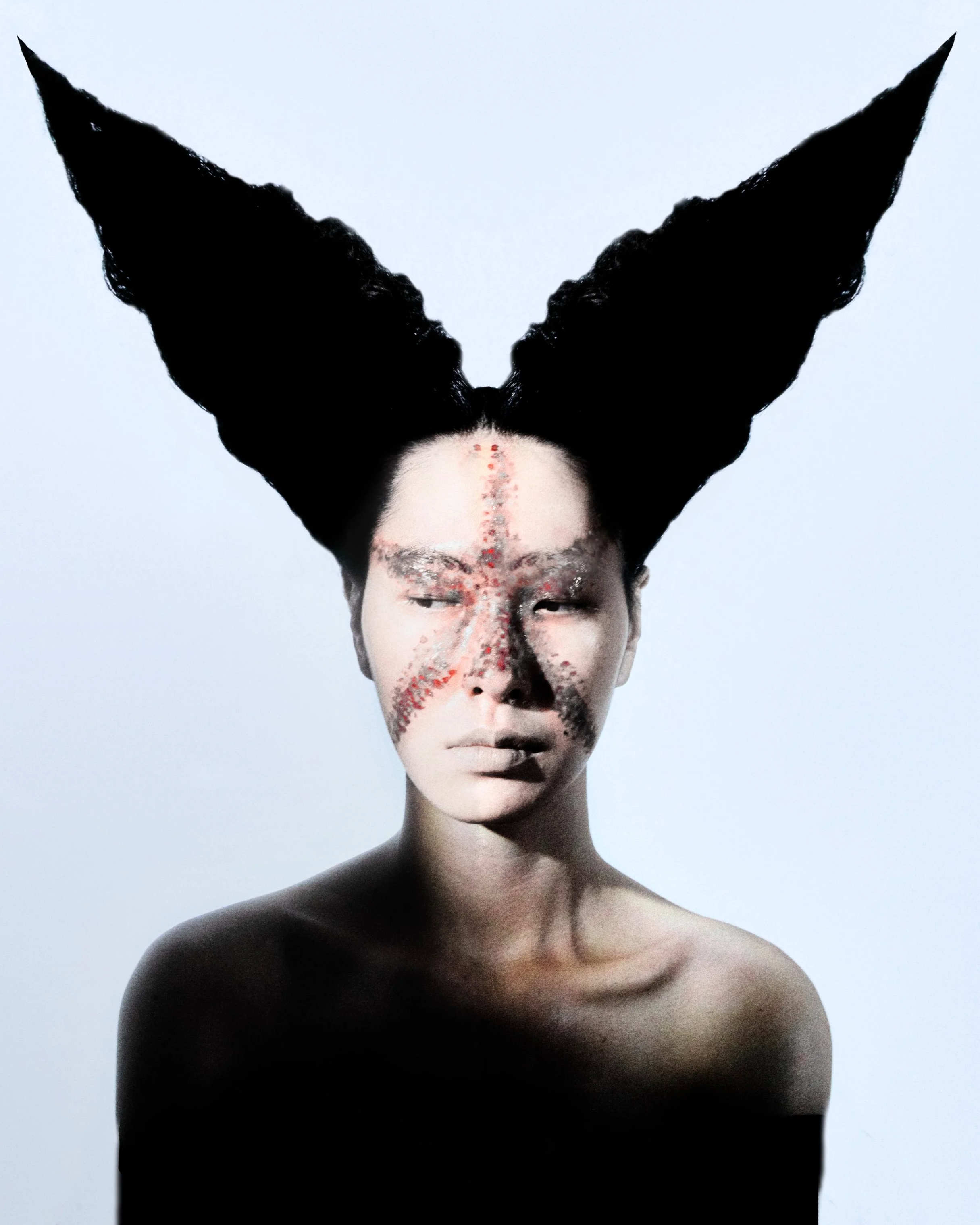A person with white face makeup and red and black painted designs on their face, with large black origami-style paper wings extending from their head, against a plain white background.