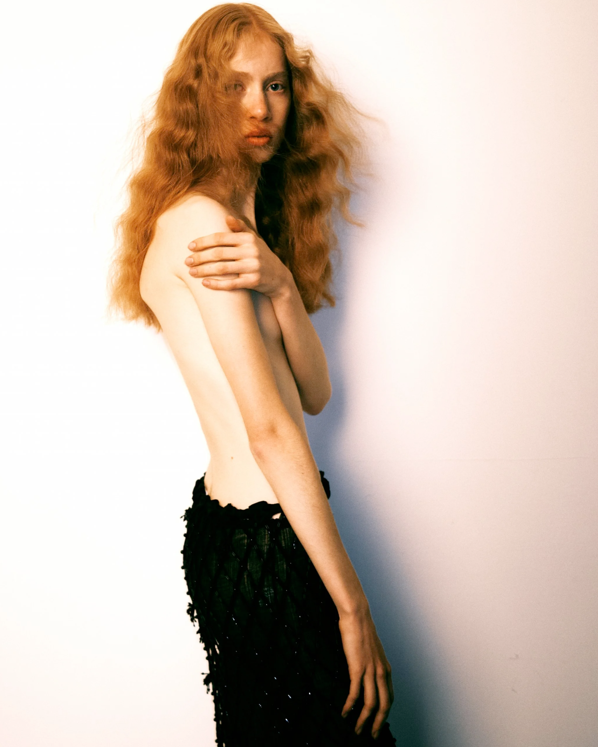 A woman with long, curly red hair standing against a plain white background, topless, with one arm crossed over her chest and the other hanging down, wearing a black lacy skirt.