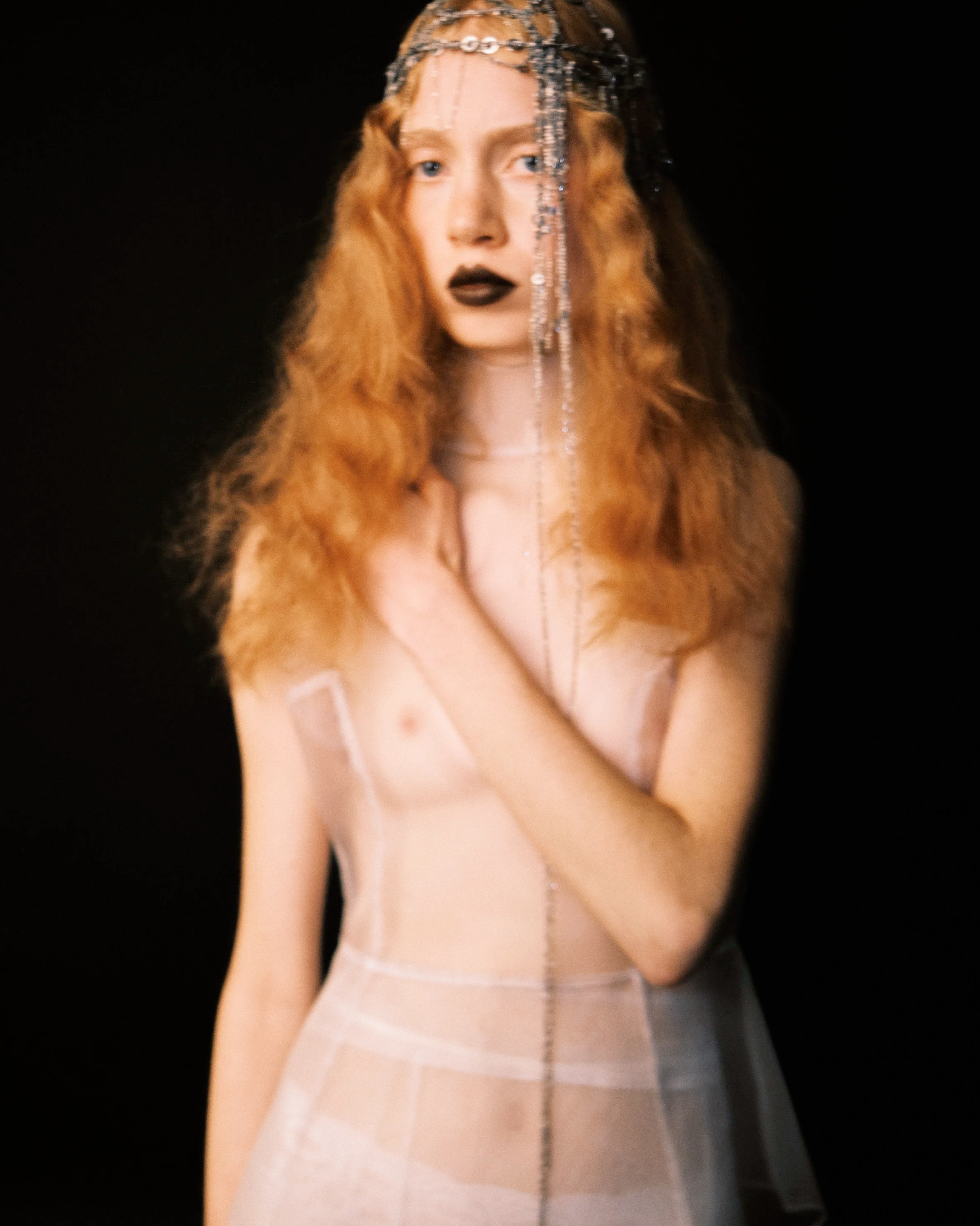 A woman with long, wavy red hair, black lipstick, and a bejeweled headpiece. She is dressed in a sheer outfit and poses against a black background.