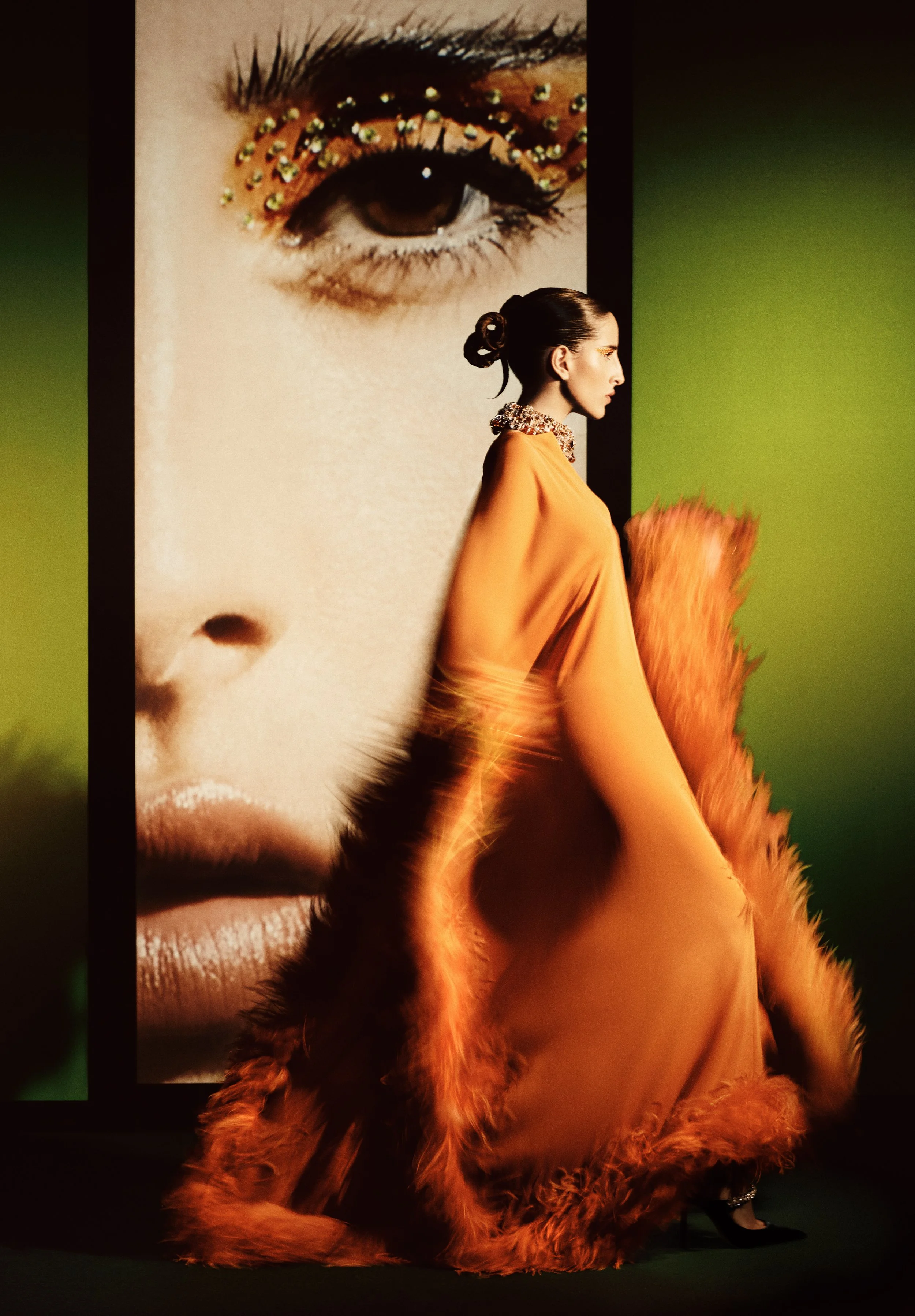 A woman with dark hair in a bun, wearing an orange gown with a floral collar, is walking past a large image of a woman's face with elaborate eye makeup and rhinestones on her eyelid.