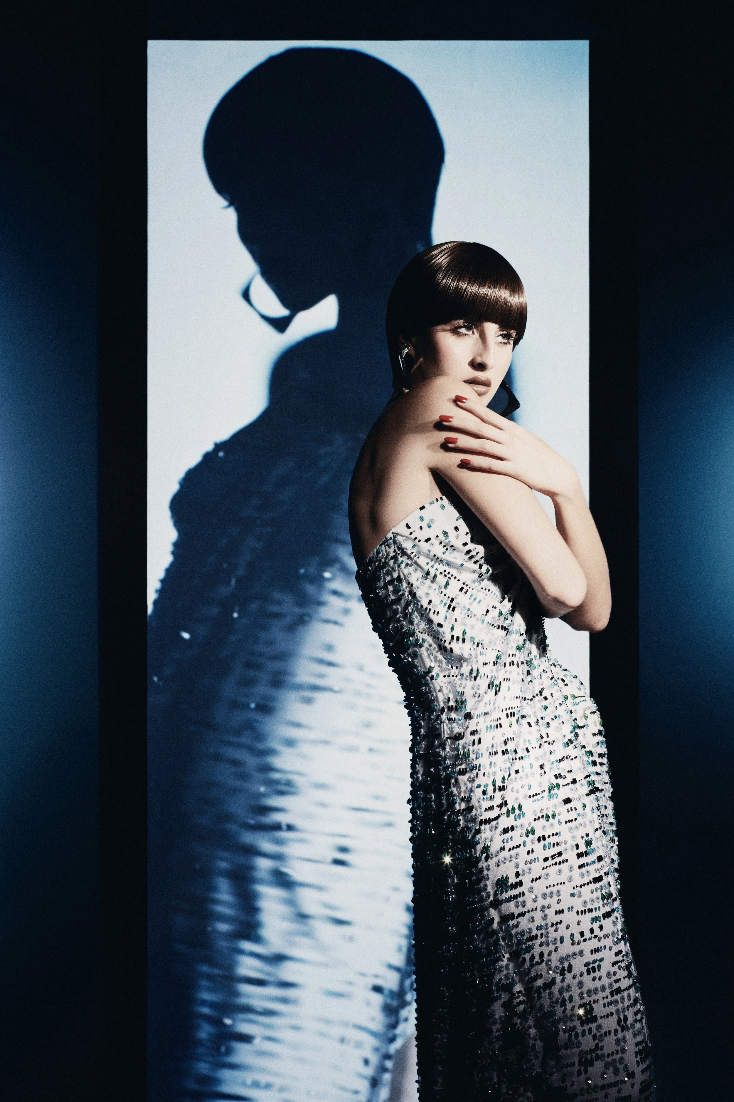 A woman with short dark hair and red lipstick posing in a shiny, sequined dress in front of a backdrop creating a shadow of her profile with a water reflection.