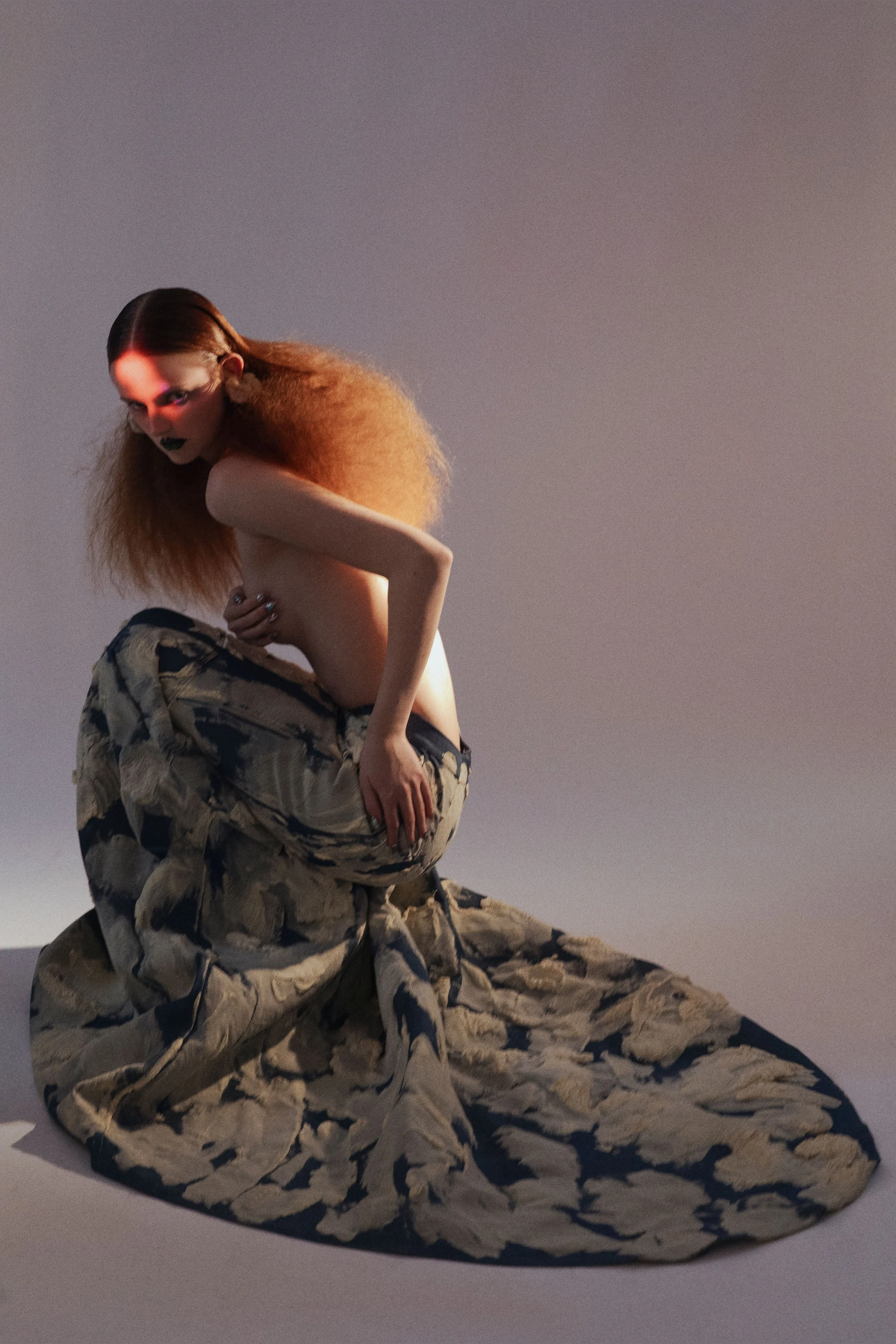 A woman with voluminous orange hair and dark makeup crouches on the floor, wearing a long, patterned skirt but no top, against a plain background.