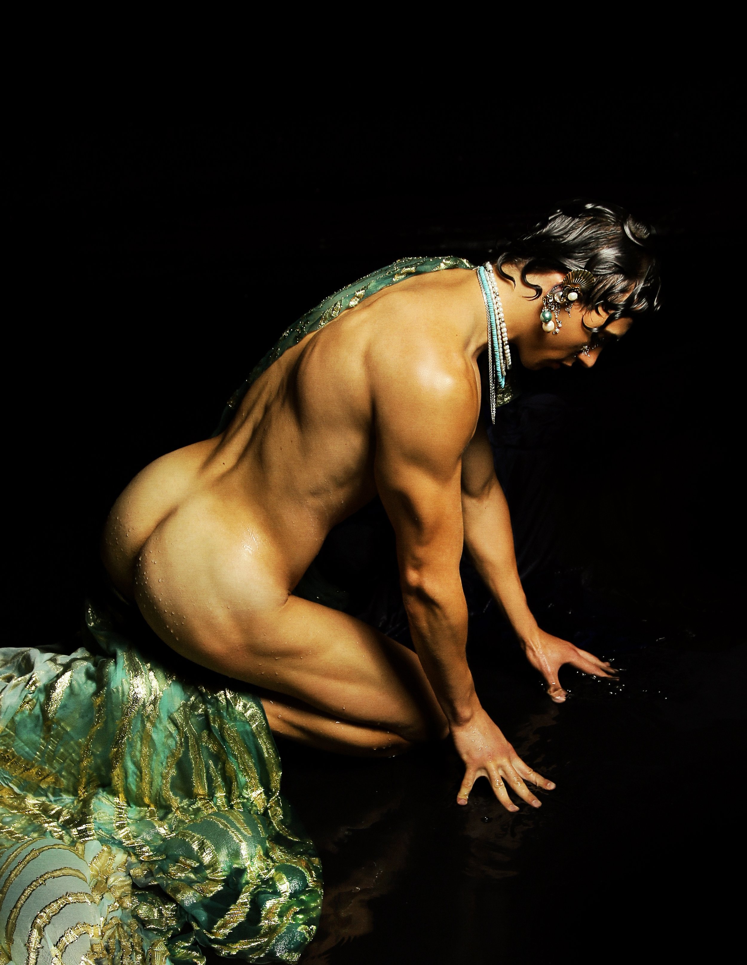 A man in a decorative outfit crouches on the ground, with his hands touching the water, against a dark background.