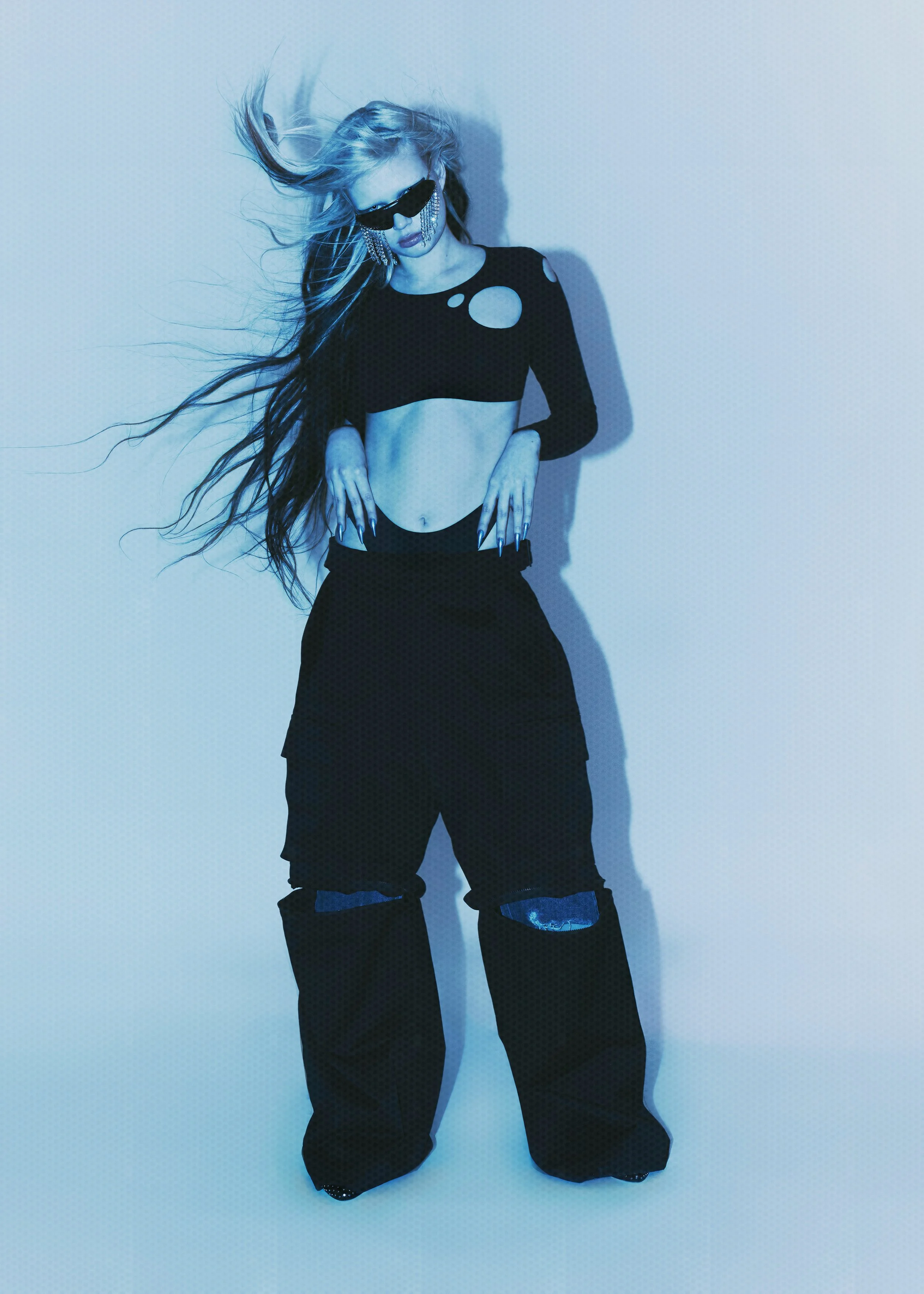 A woman with long, flowing hair wearing black clothing with cutouts, oversized pants with ripped knees, and sunglasses, standing against a plain background with a blue tint.