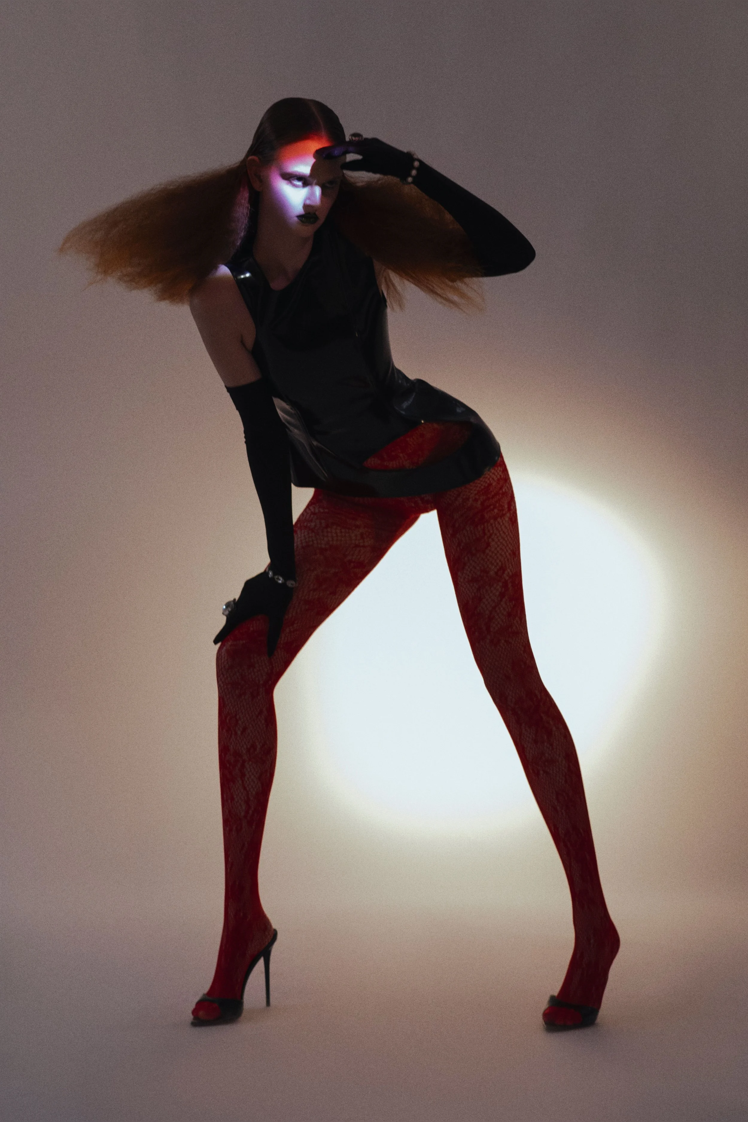 Fashion model with styled hair, dark makeup, wearing black sleeveless top, red patterned tights, black high heels, stands in a dramatic pose against a plain background with a bright light behind