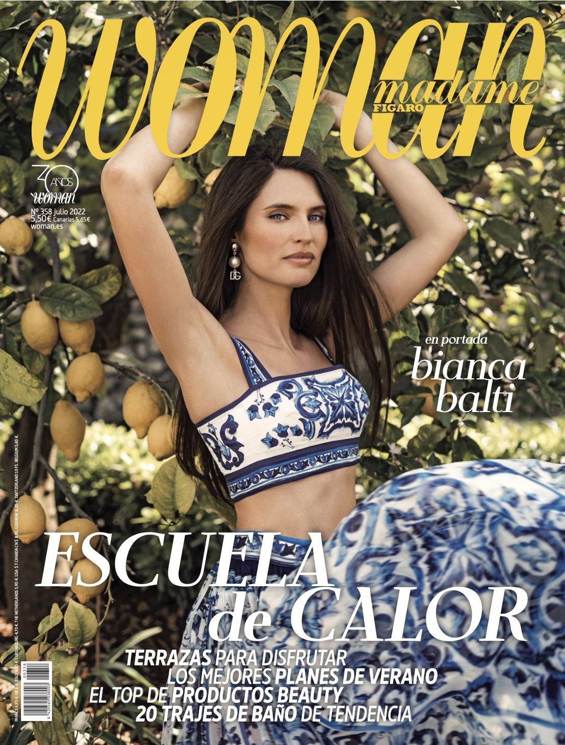 A woman wearing a blue and white patterned crop top and matching skirt posing outdoors among lemon trees with ripe lemons, with her hands raised above her head.
