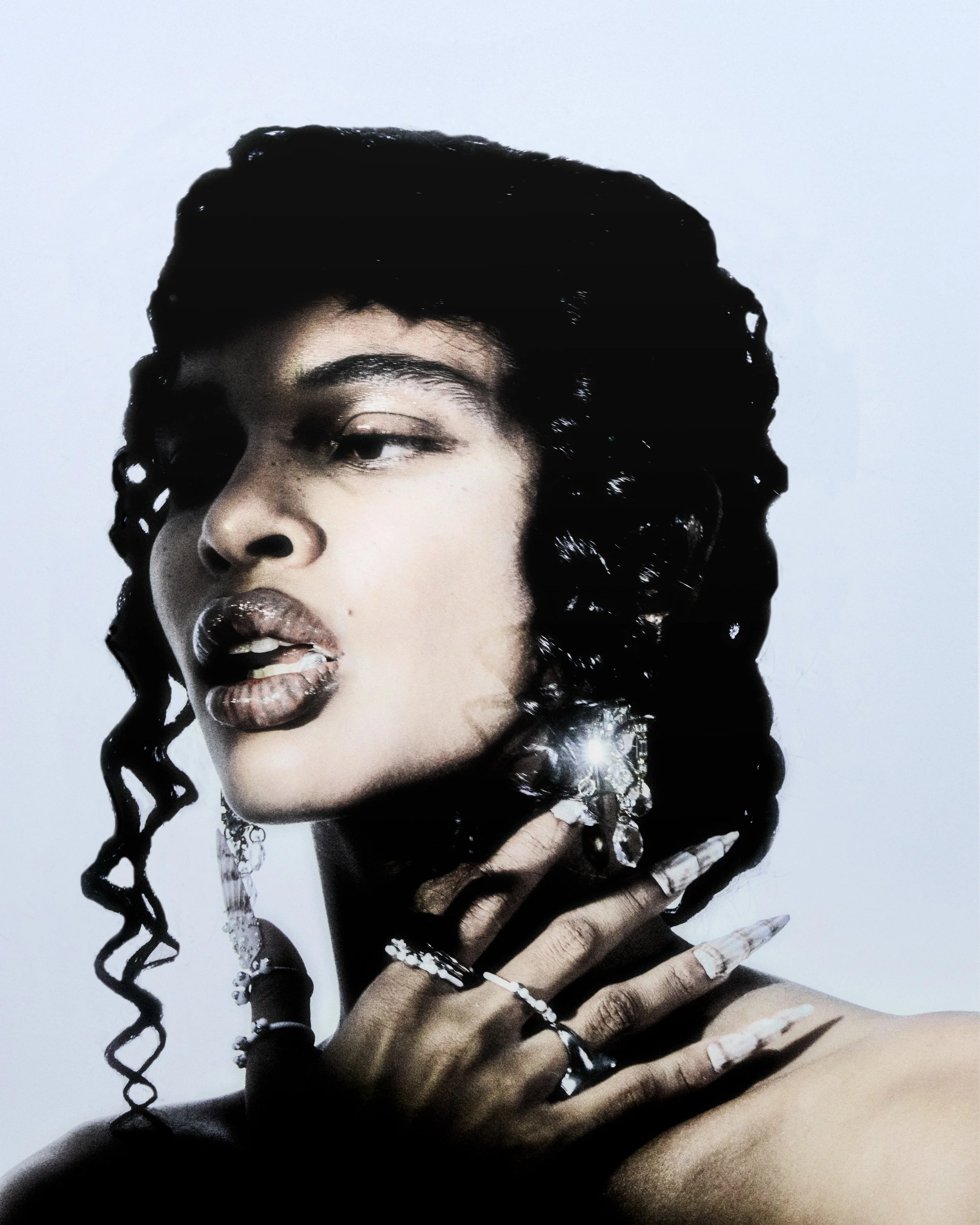Portrait of a woman with dark curly hair, wearing multiple rings, earrings, and necklaces, with makeup and a confident expression.