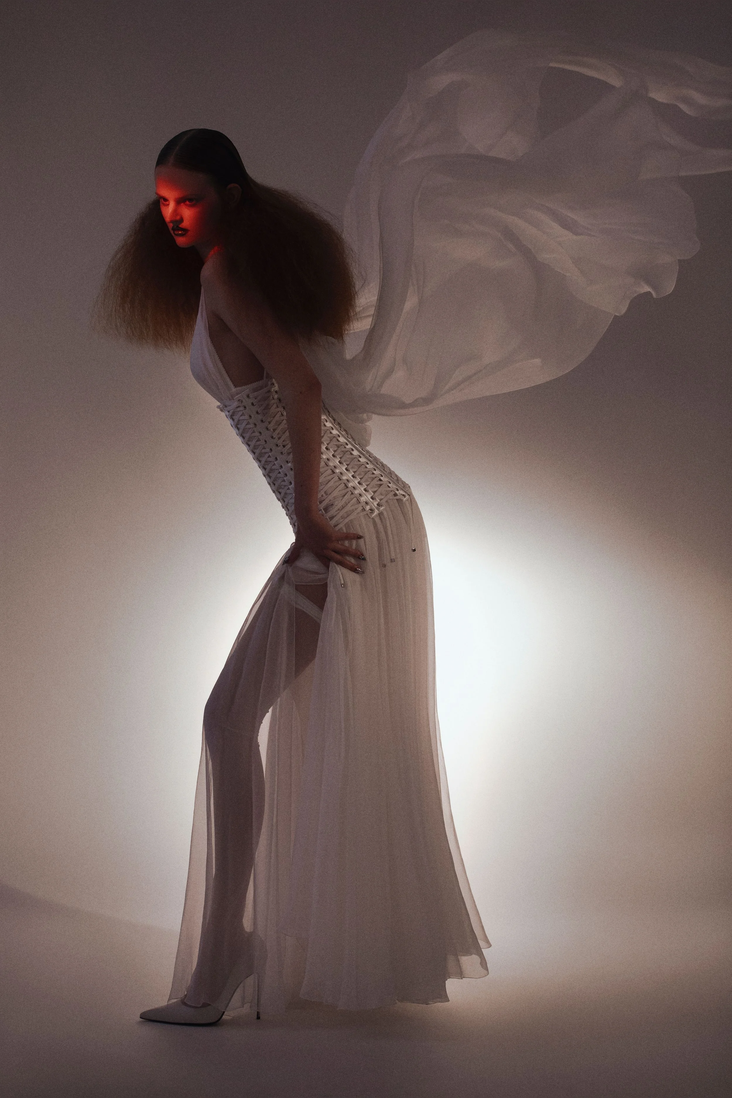 A woman in a long, flowing white dress with wide-brimmed heels and styled hair, posing against a neutral background with lighting that creates shadow and highlights.