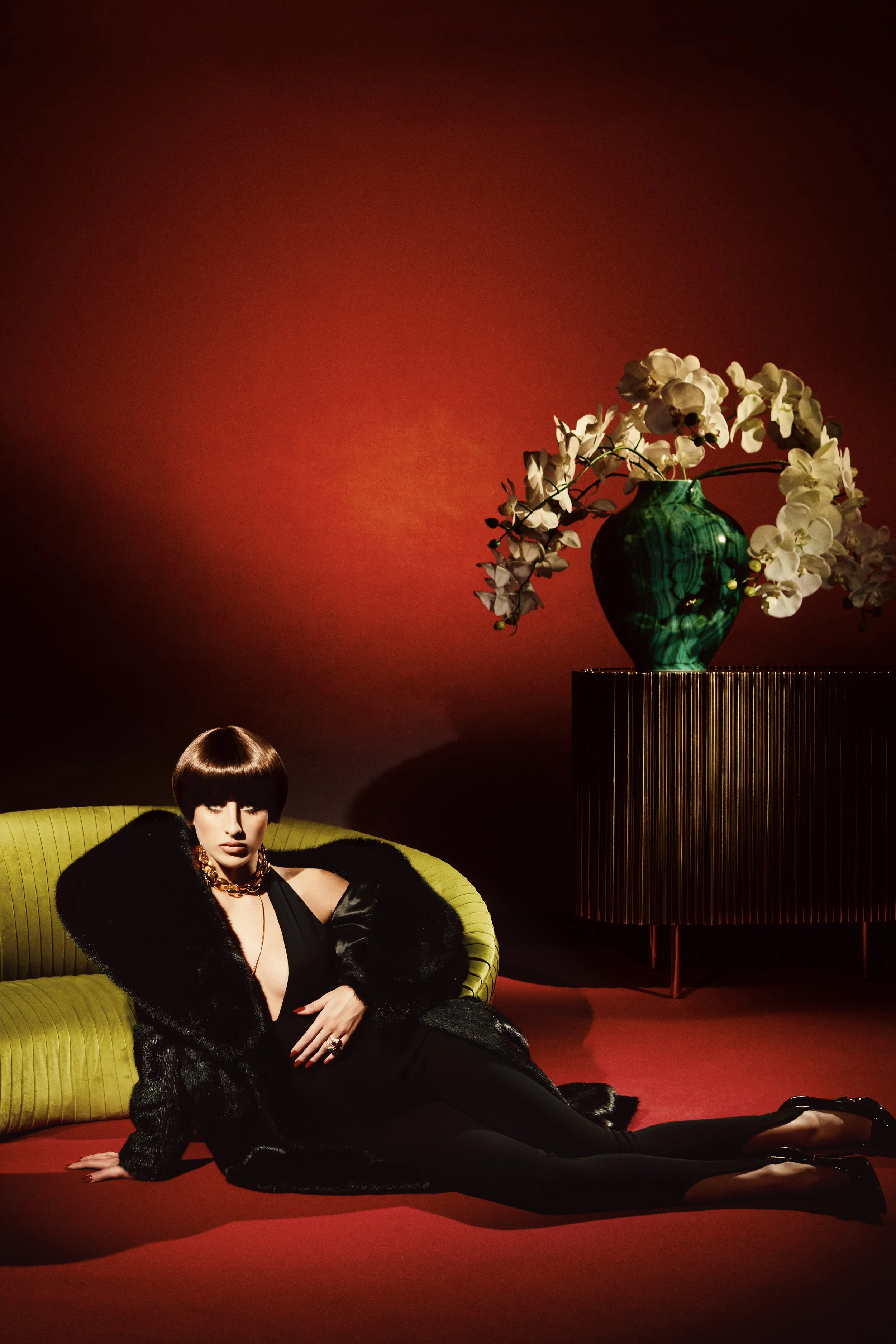 A woman with a short dark bob haircut and wearing a black dress, black tights, and heels, is reclining on a bright yellow velvet sofa. She has a black fur coat draped over her shoulders, jewelry, and makeup, with a serious expression. Behind her, the