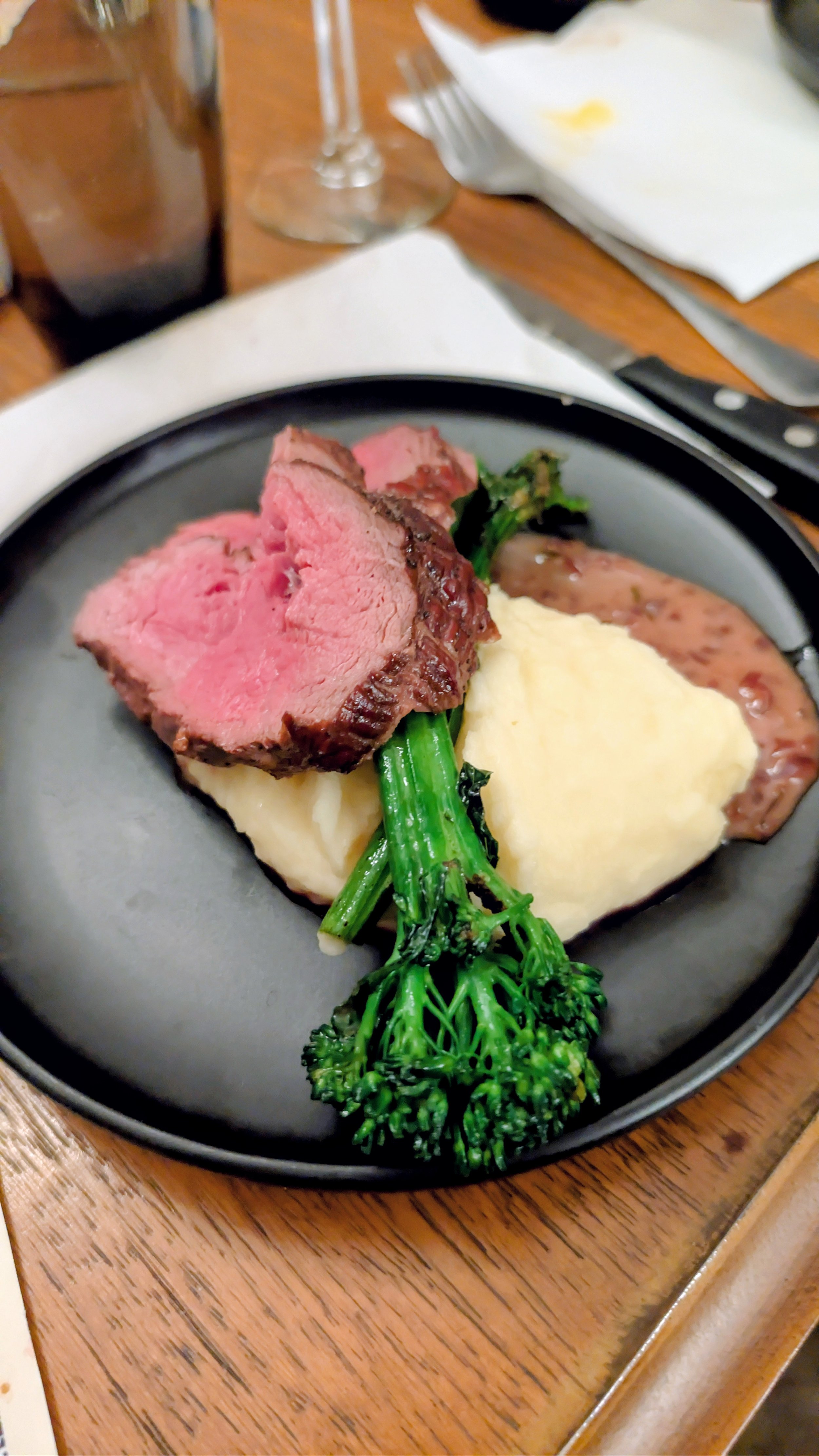A plated dinner with slices of medium-rare beef tenderloin, mashed potatoes, broccolini, and gravy, served on a black plate.