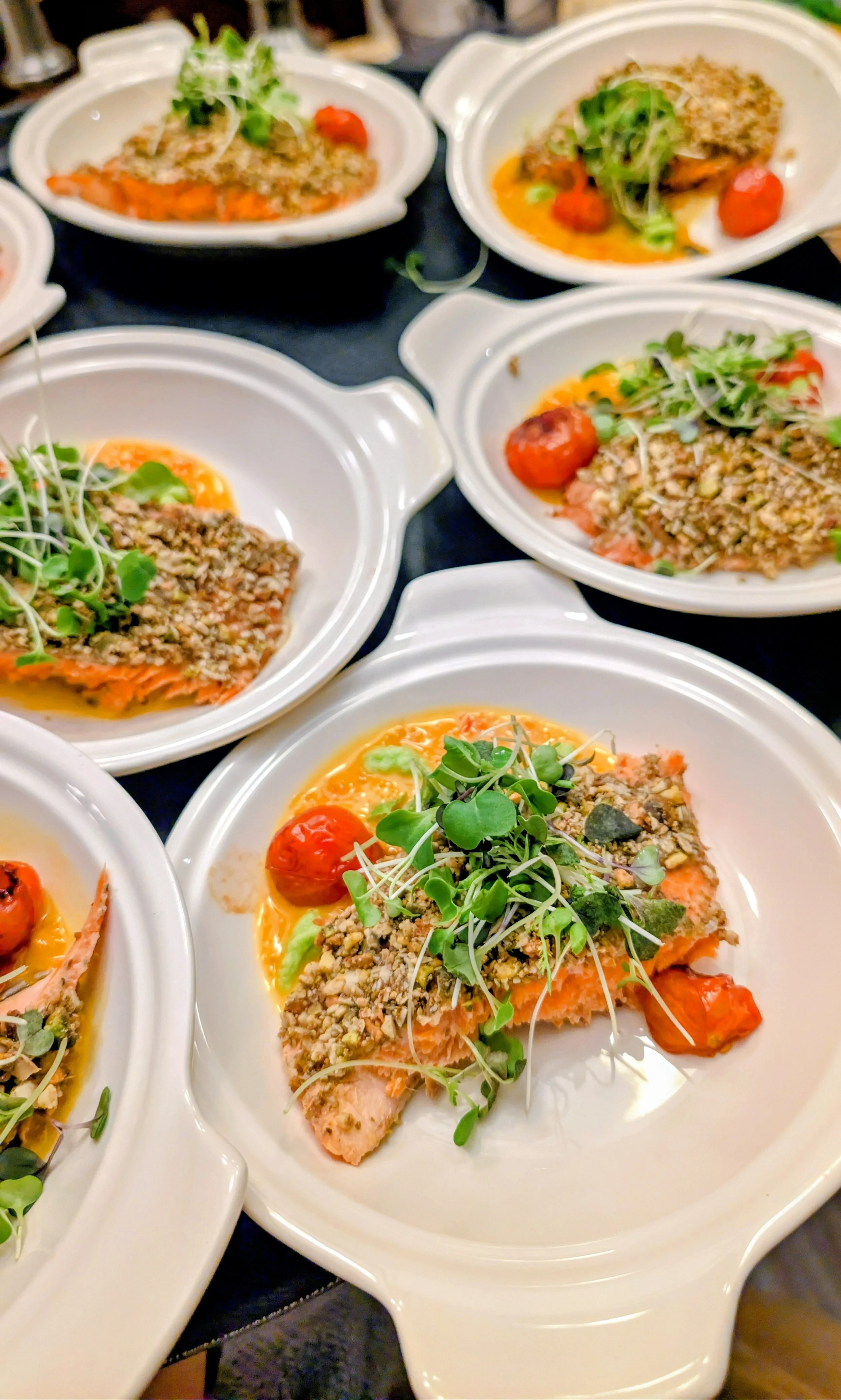 Plates of cooked salmon topped with microgreens, chopped nuts, cherry tomatoes, and sauce.