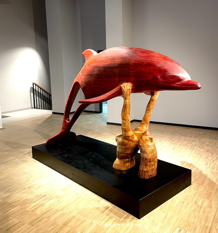 Eine große Skulptur eines fliegenden Vogels, die auf zwei Beinen steht, im Inneren eines Museums oder Kunsthalle. Die Skulptur ist hauptsächlich rot mit holzähnlichen Texturen an den Beinen und steht auf einem schwarzen Podest.