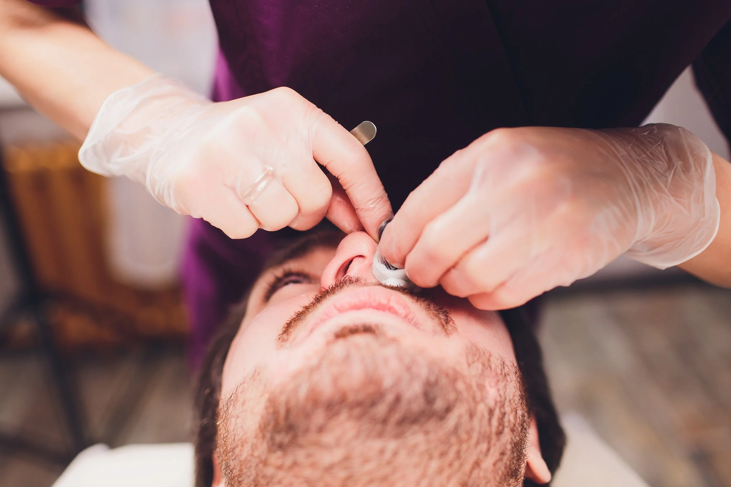 Professional technician using hard wax to remove hair from client’s nose, wearing gloves