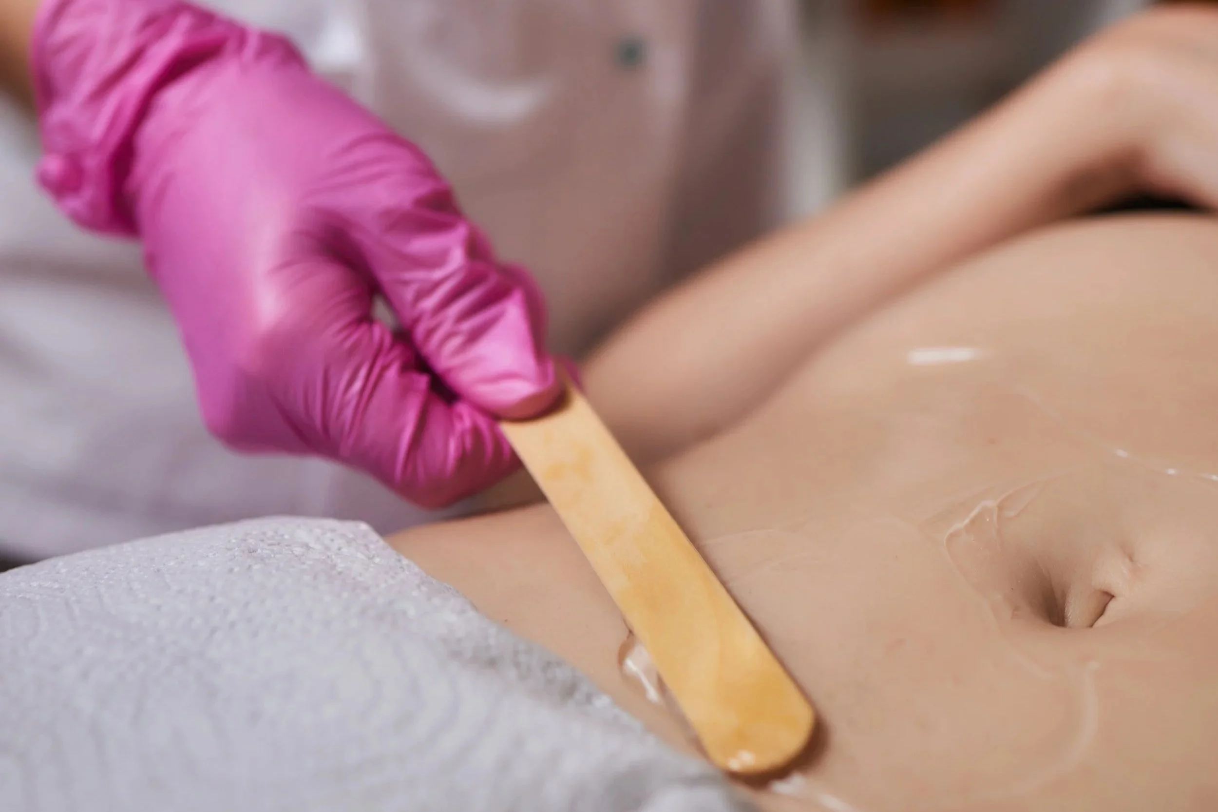 A technician in pink gloves is applying wax to a client’s abdomen using a wooden spatula during a waxing procedure.