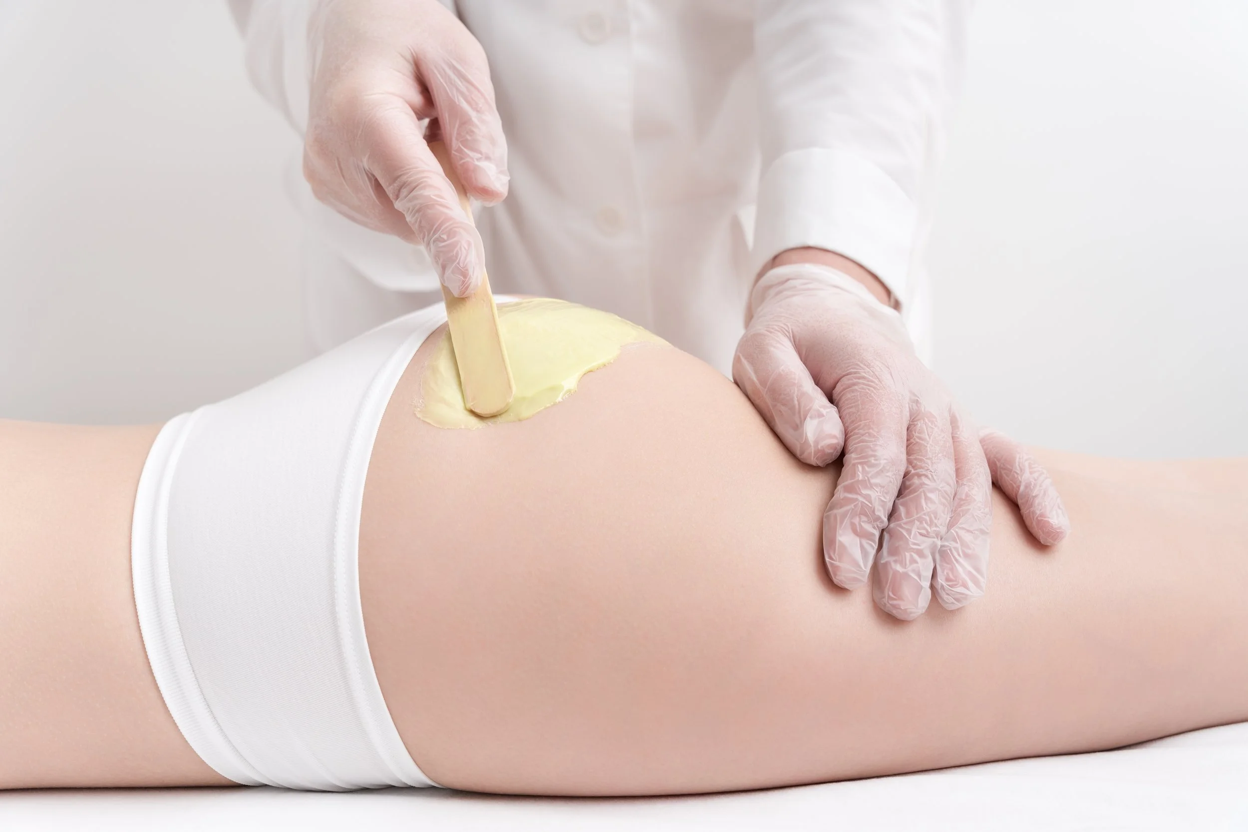 Esthetician applies soft wax to glutes using a wooden spatula, wearing disposable gloves.
