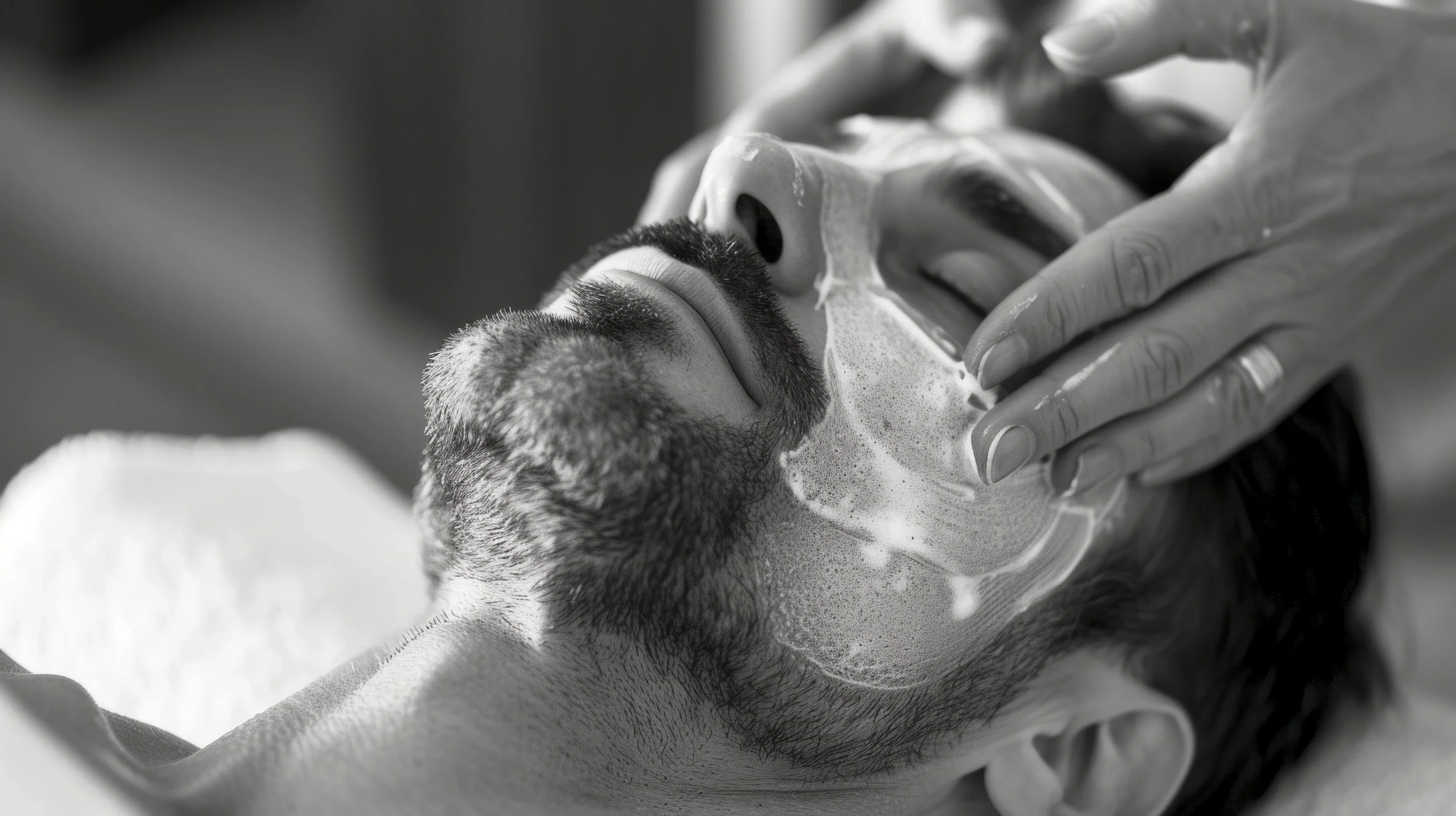 Close-up of a bearded man receiving a facial treatment while an esthetician massages cleanser onto his face