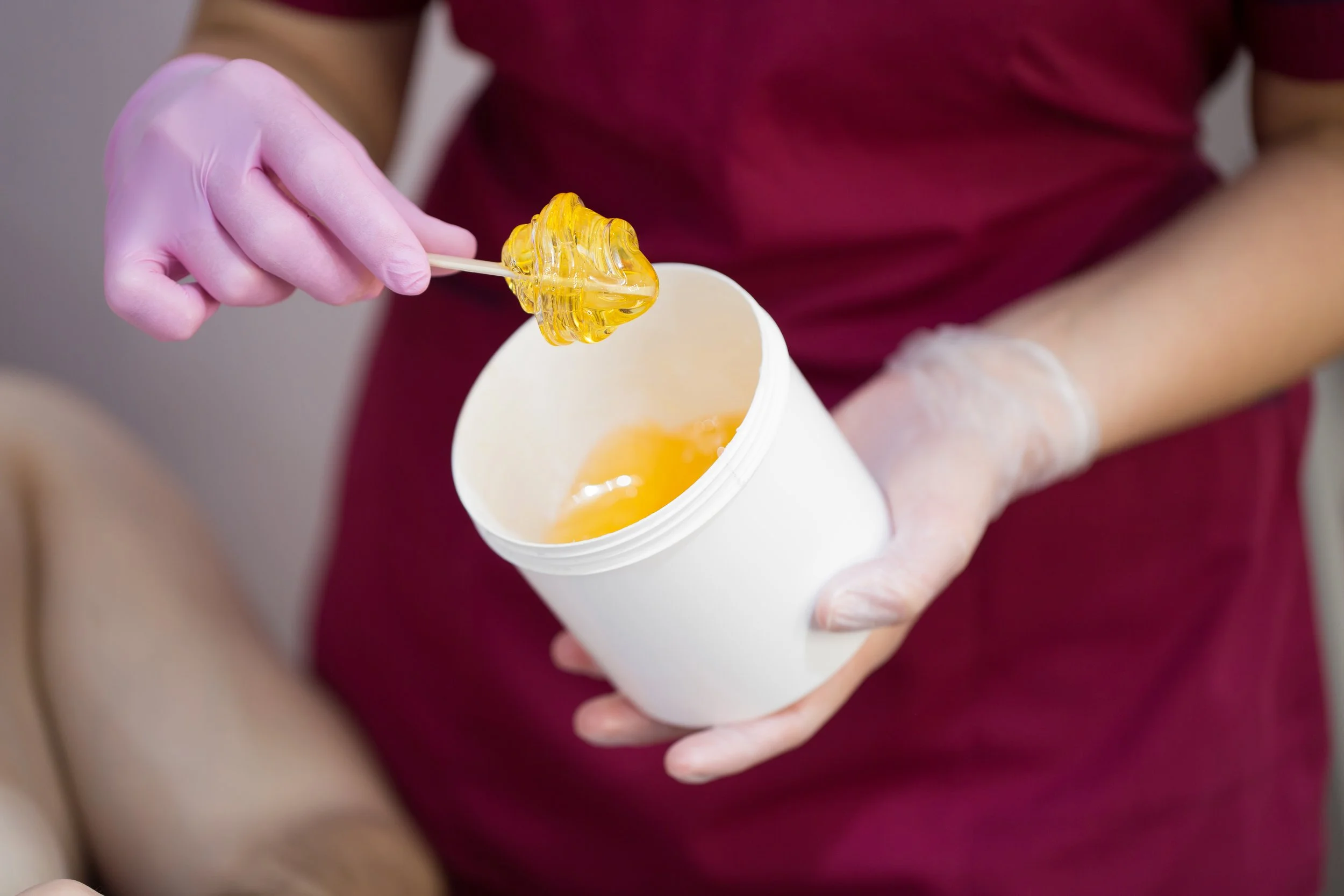Hand holding a wax pot with wooden spatula for hair removal.