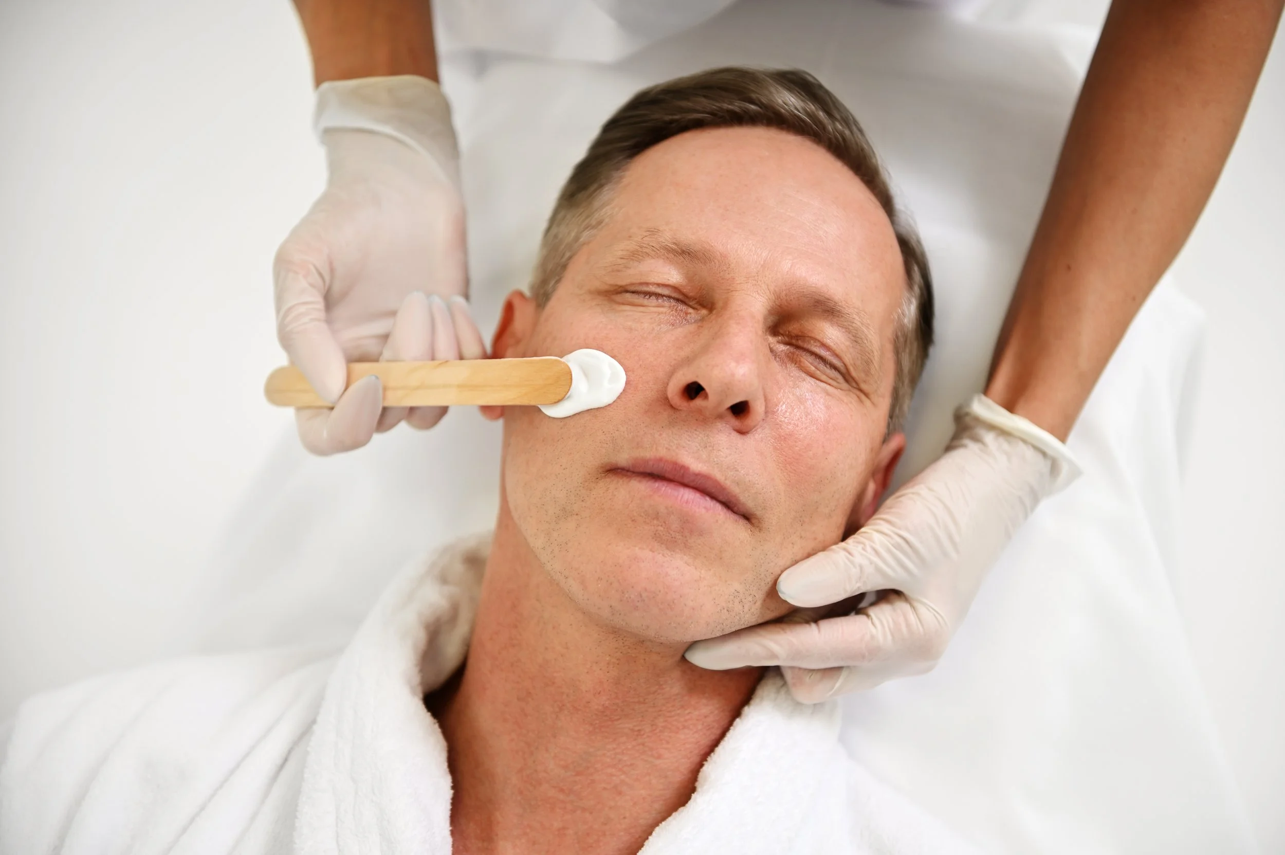A male client receives a facial waxing treatment. The specialist in medical gloves is applying wax to the client’s face using a wooden spatula. The client is lying down with his eyes closed.
