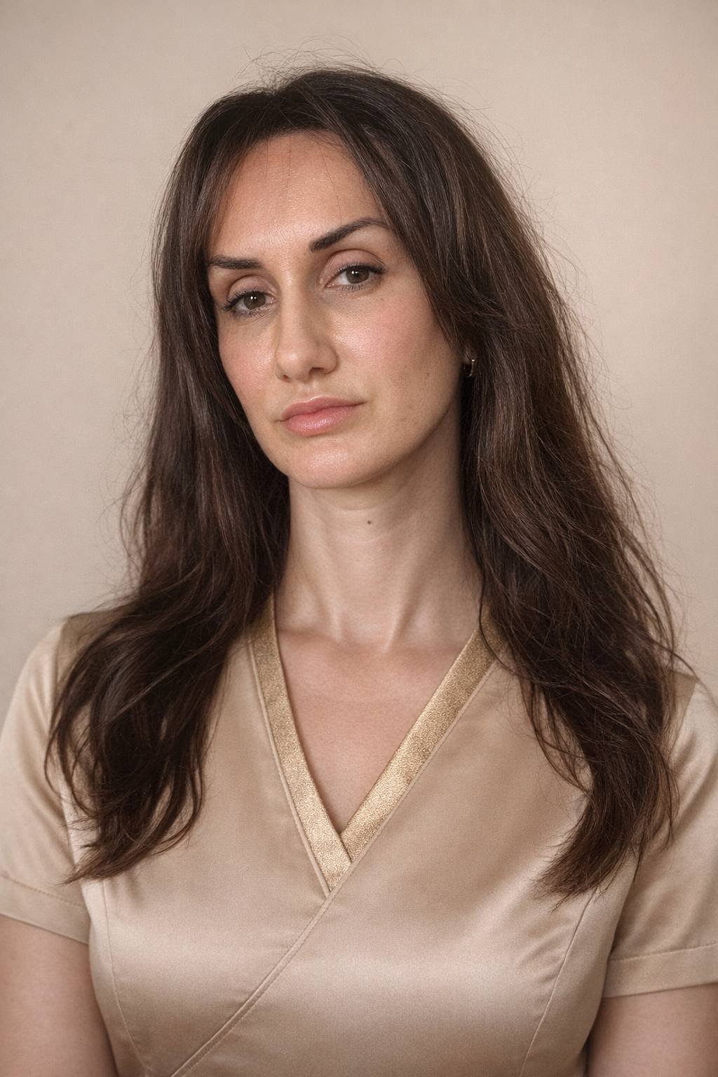 Portrait of a woman with  hair, blue eyes, wearing a white blazer and a delicate cross necklace, against a plain light background.