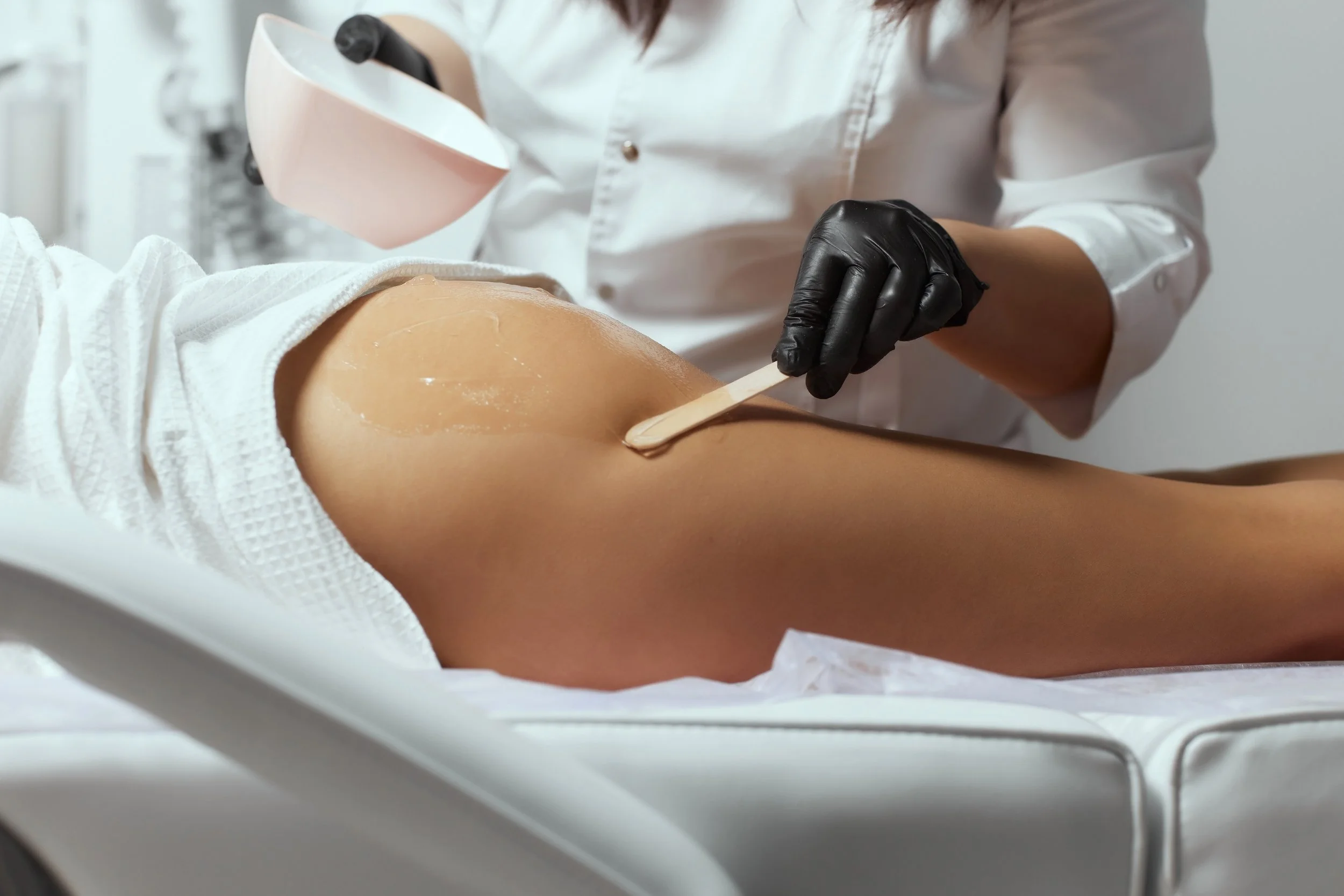 Esthetician applies soft wax to glutes using a wooden spatula and black gloves.