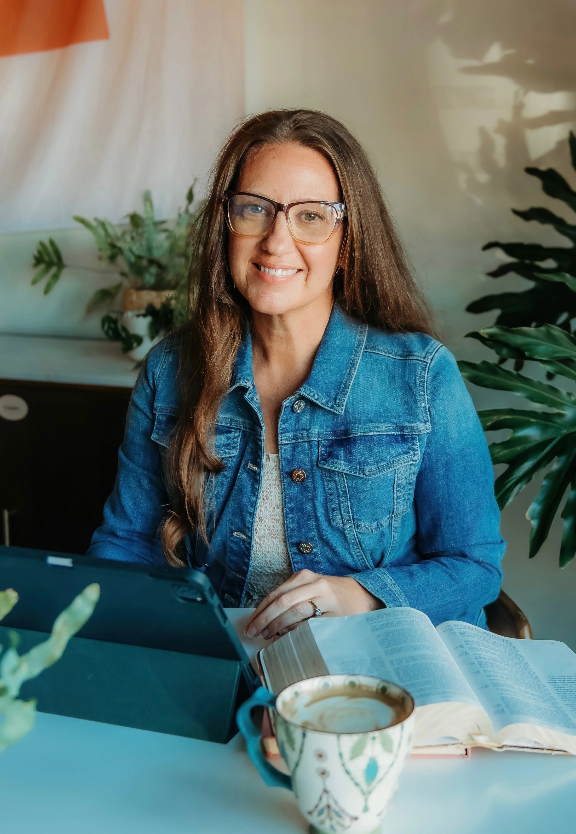 A woman with long brown hair, glasses, and a denim jacket is sitting at a table with an open book, a tablet, and a colorful coffee mug. She is smiling and looking at the camera, with green plants and a light-colored wall in the background.