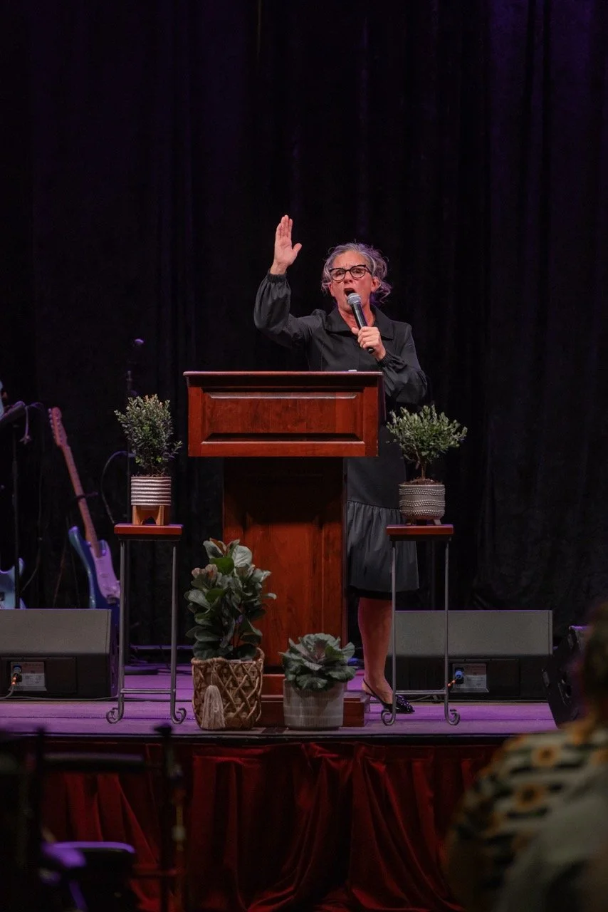 A woman with gray hair and glasses speaking passionately at a podium on a stage, with plants and musical instruments visible around her.