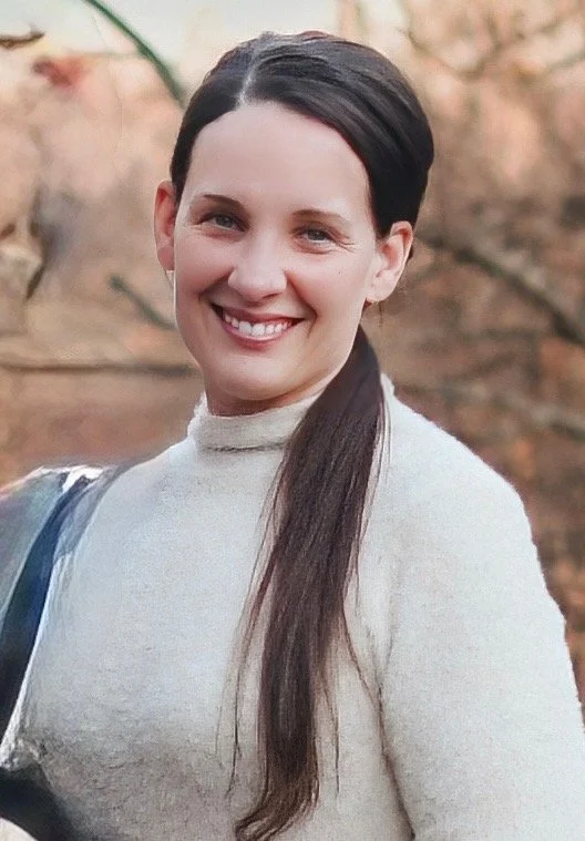 A woman with long dark hair wearing a light-colored turtleneck sweater, smiling outdoors with a blurred background of trees and autumn leaves.