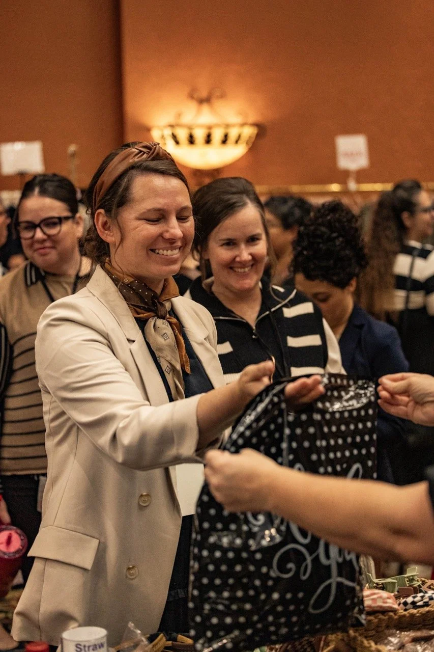 Smiling woman in a beige blazer shopping at an indoor market or fair, surrounded by other shoppers.