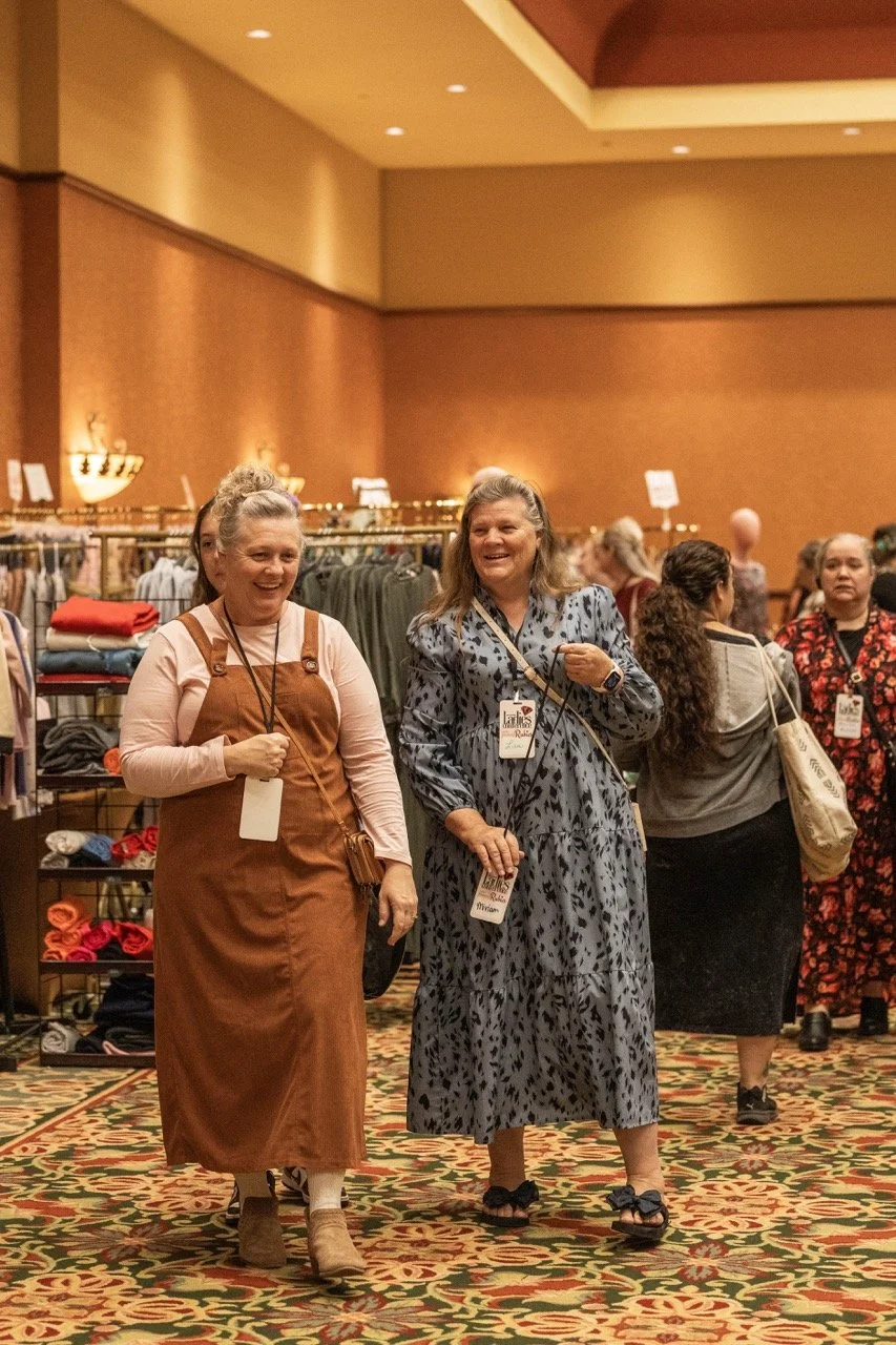 Two women walking and chatting at an indoor event, surrounded by other attendees and clothing racks.